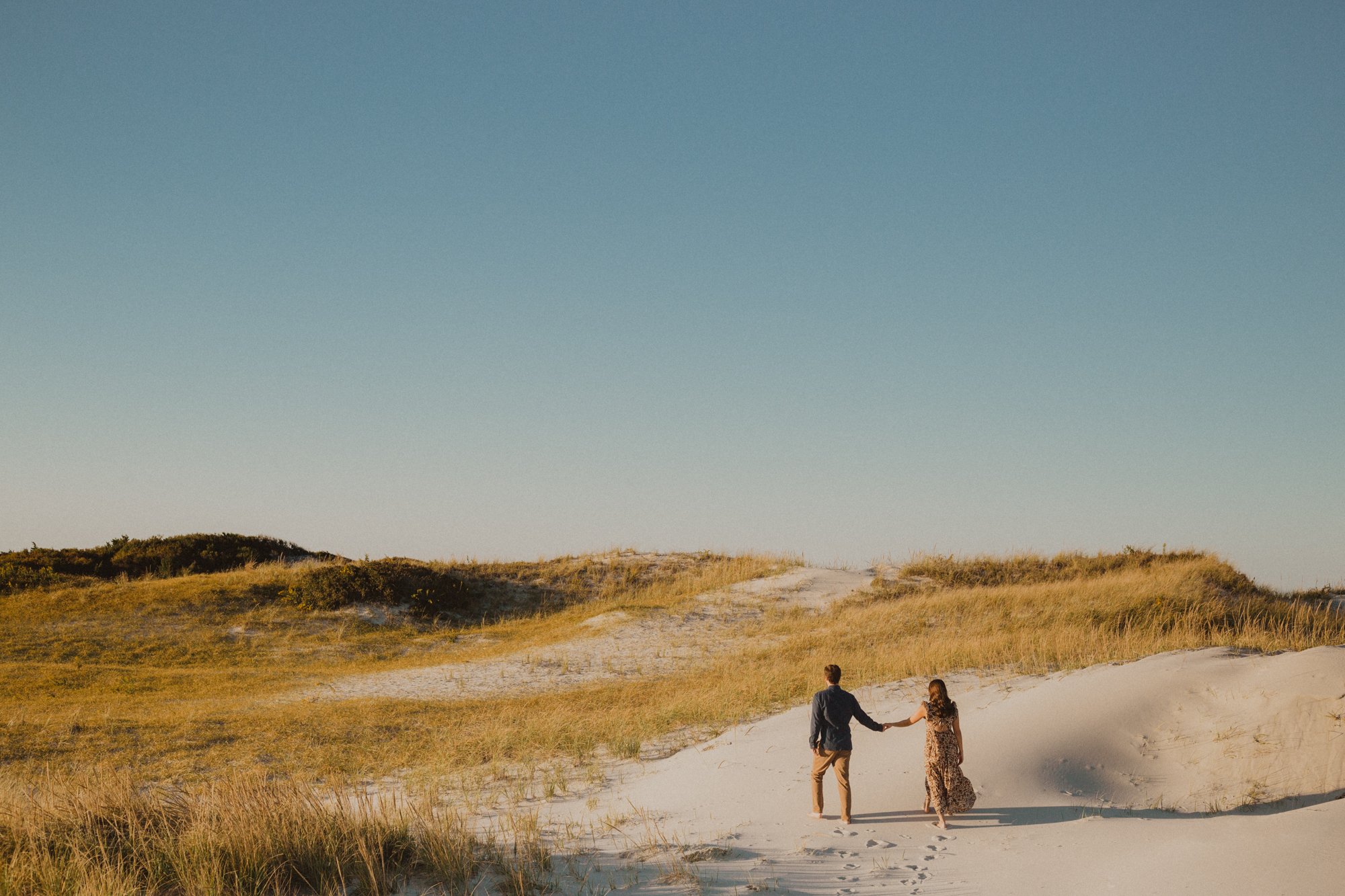 Wide angle photo of a couple walking in sand dunes, holding hands in natural light at sunset