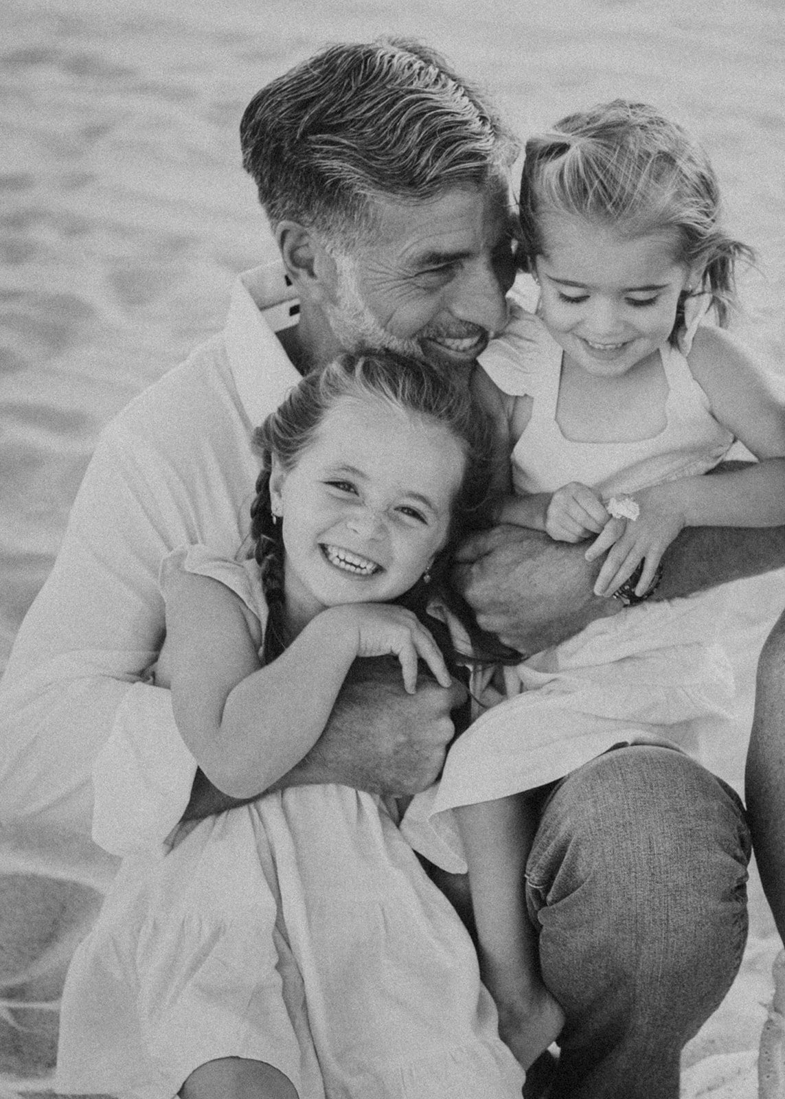 Father sat in sand on the beach, holding two young grandaughters as they smile at camera.