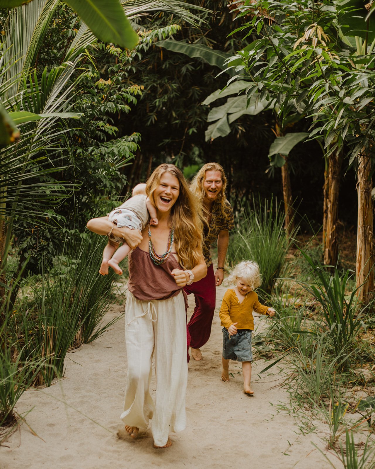 Woman carrying newborn baby over shoulder and laughing while walking through green jungle, with man and young boy catching up behind her.