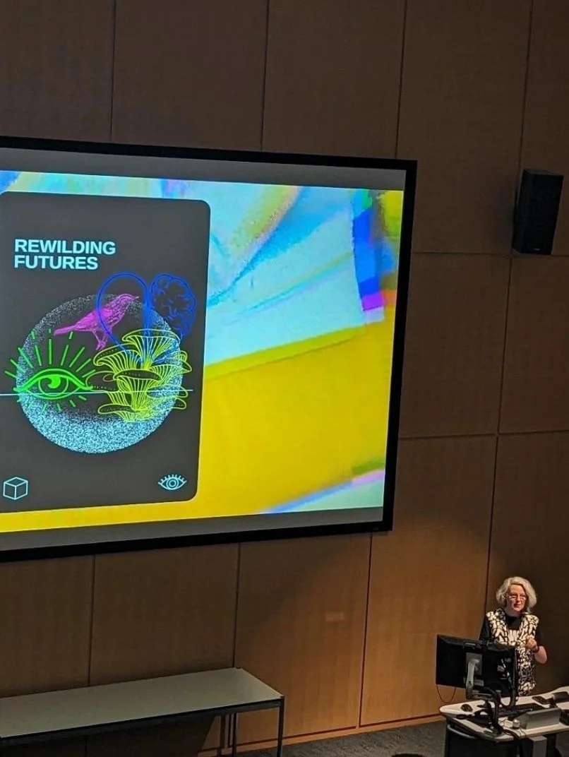 A futurist speaker giving a presentation with the title 'Rewilding Futures' displayed on a large screen. The screen shows colorful graphics featuring abstract shapes and eyes. The speaker is standing at a podium in a lecture hall.