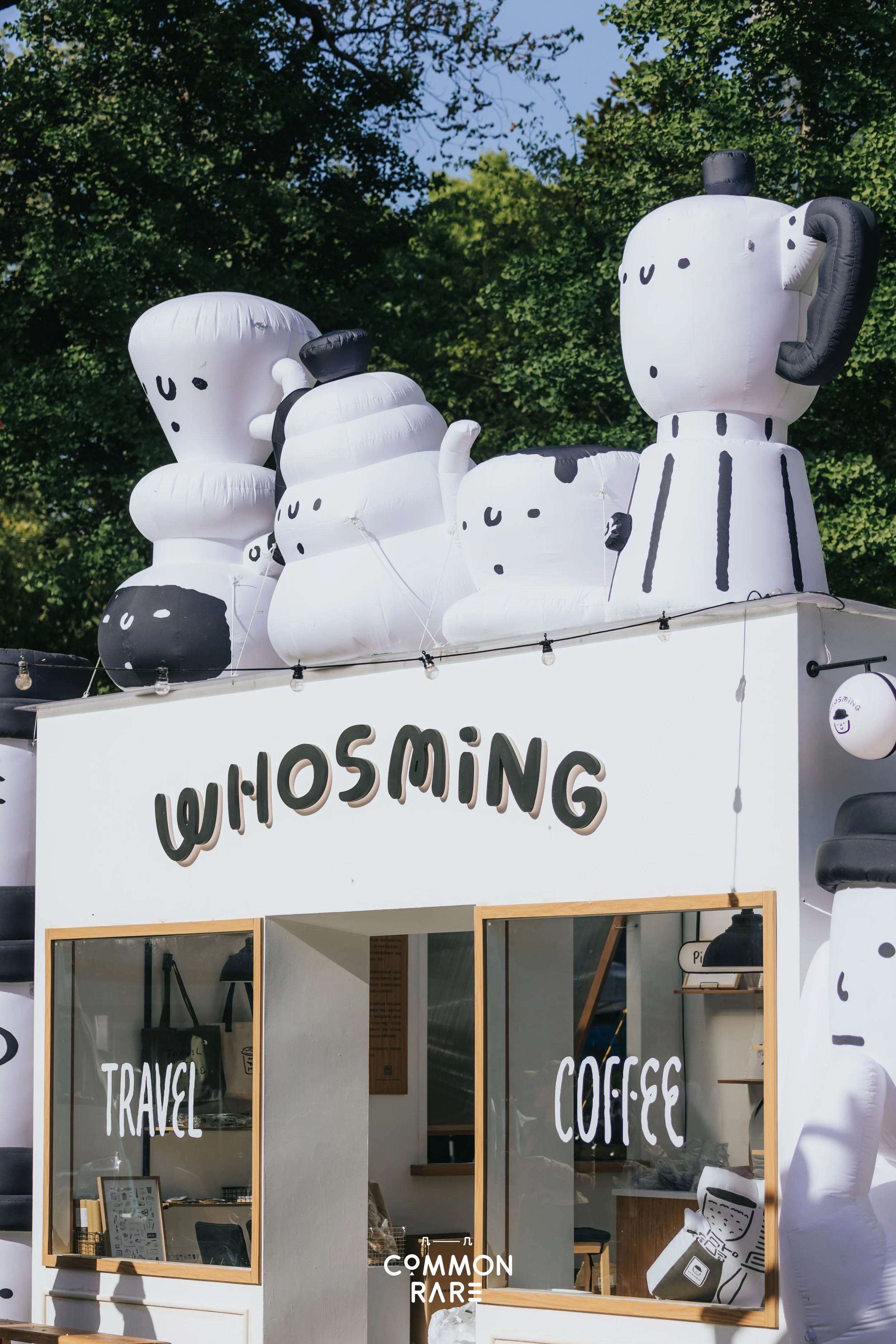Inflatable cartoon coffee mugs on top of a cafe stand, with a sign reading "WIHOUSING" and glass windows displaying words "TRAVEL" and "COFFEE," set against a background of green trees and a blue sky.
