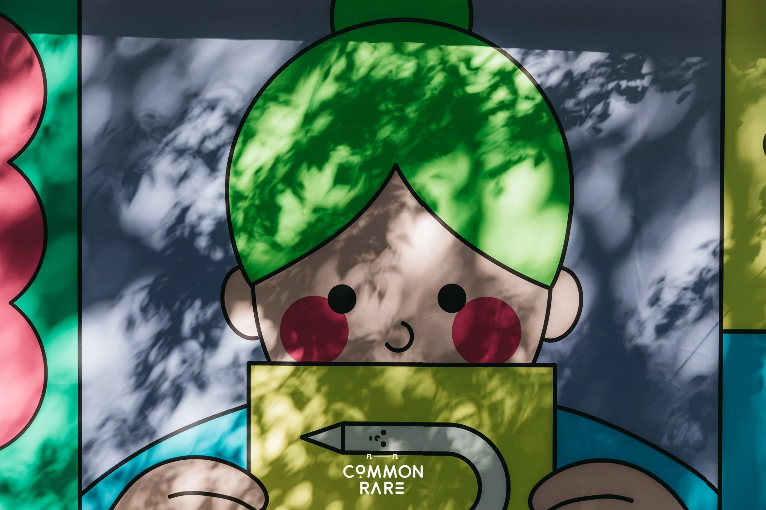 Colorful poster with a cartoon elf face with green hair, red cheeks, and a white face. The elf is smiling. The background has a pattern of clouds and shadows. Text at the bottom says "COMMON RARE" with a pencil icon above it.