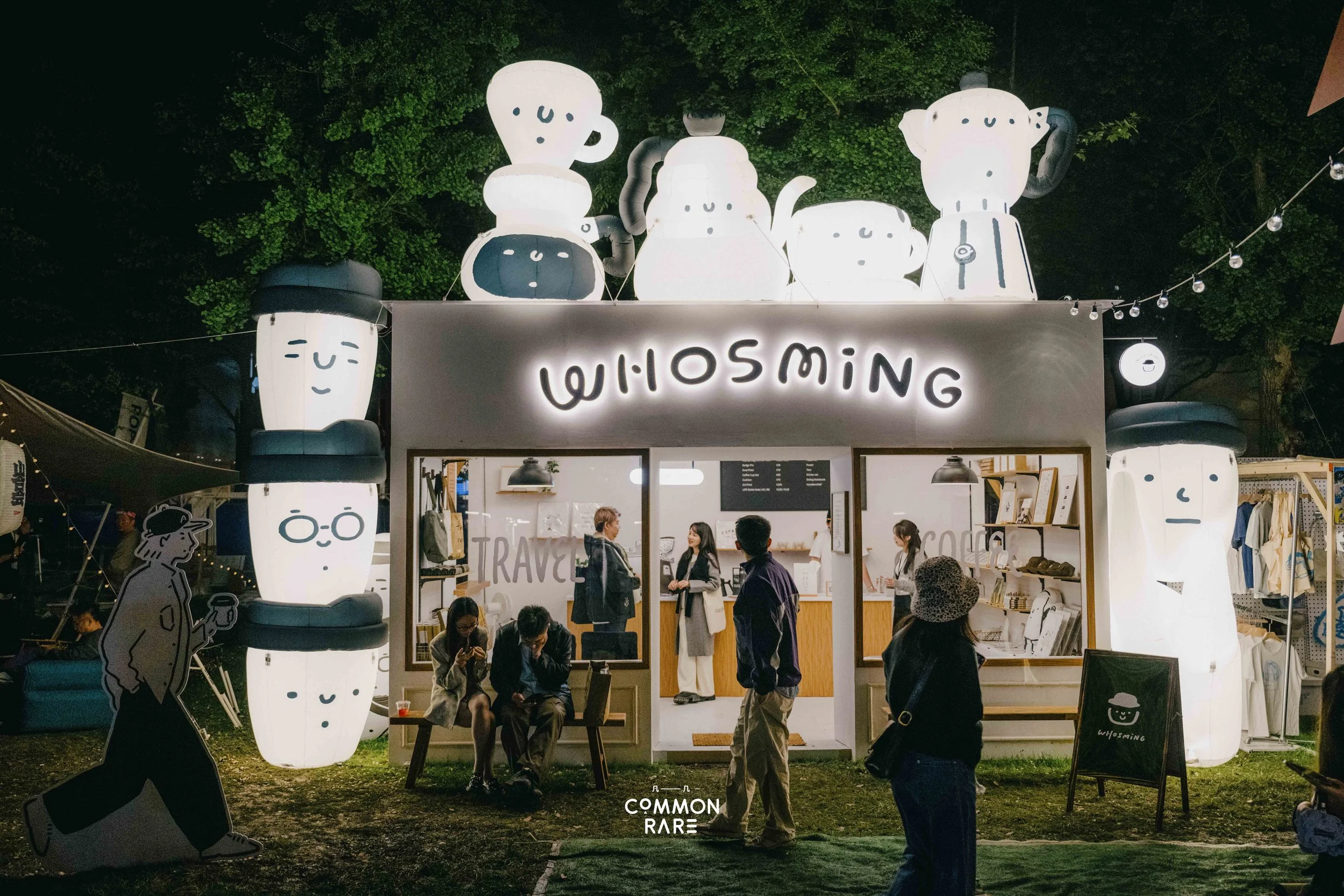 Night scene of a food or product stall with a whimsical design featuring illuminated characters resembling coffee cups and teapots with smiling faces, string lights, and people interacting in front of it.