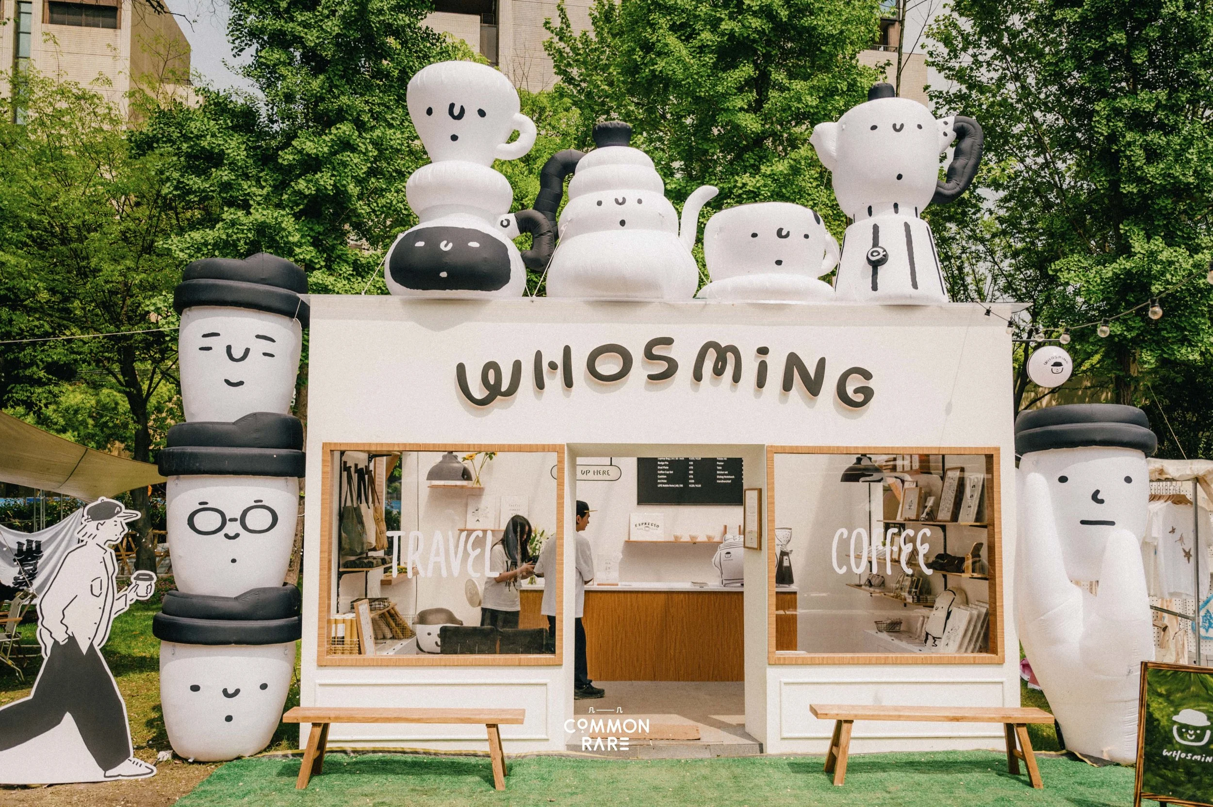 Inflatable sculptures of smiling coffee cups and teapots with black and white faces, arranged above a white booth with the word 'WISDOM' on it, set outdoors among green trees.