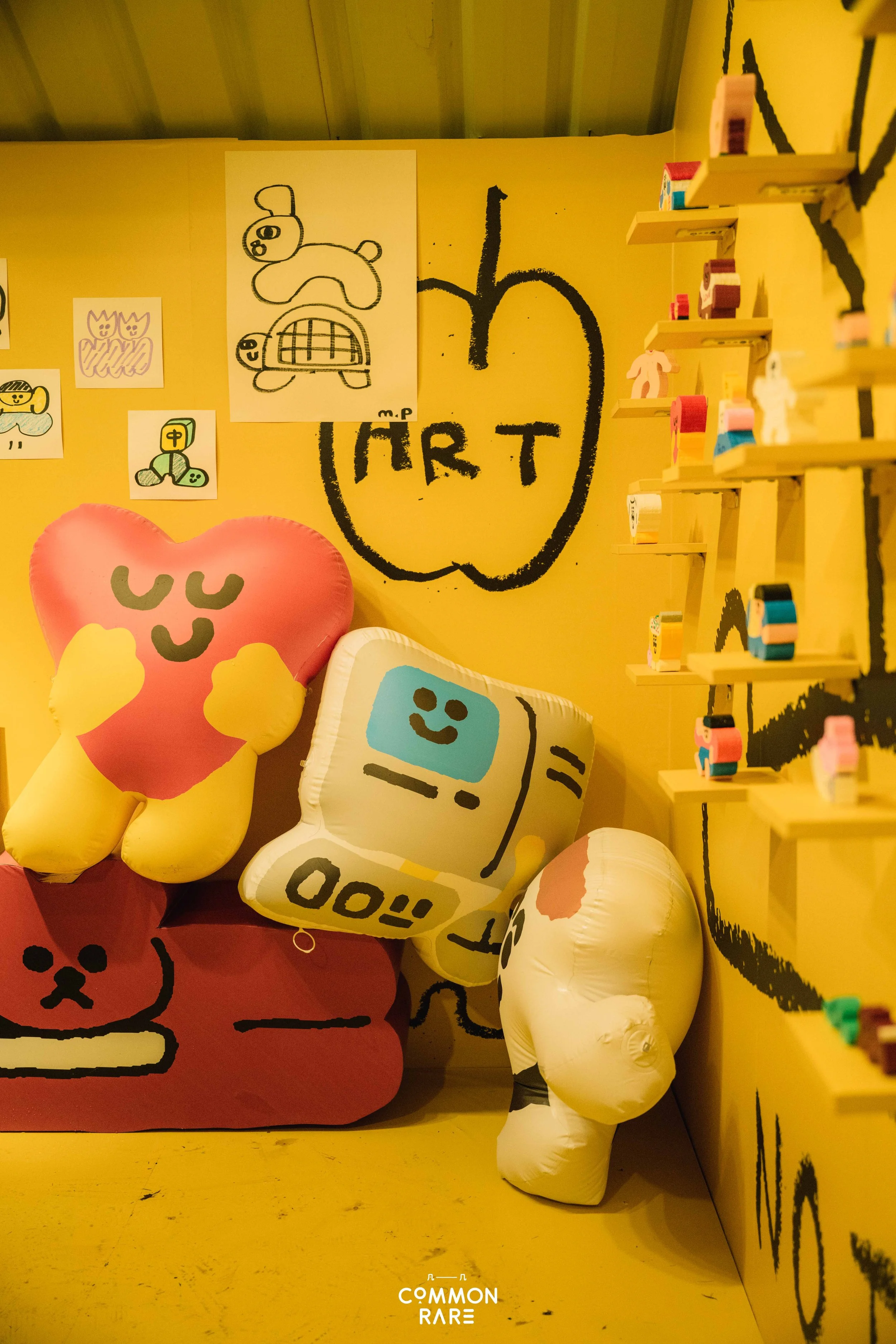 Colorful art display with plush toys, drawings, and shelves on a yellow wall.