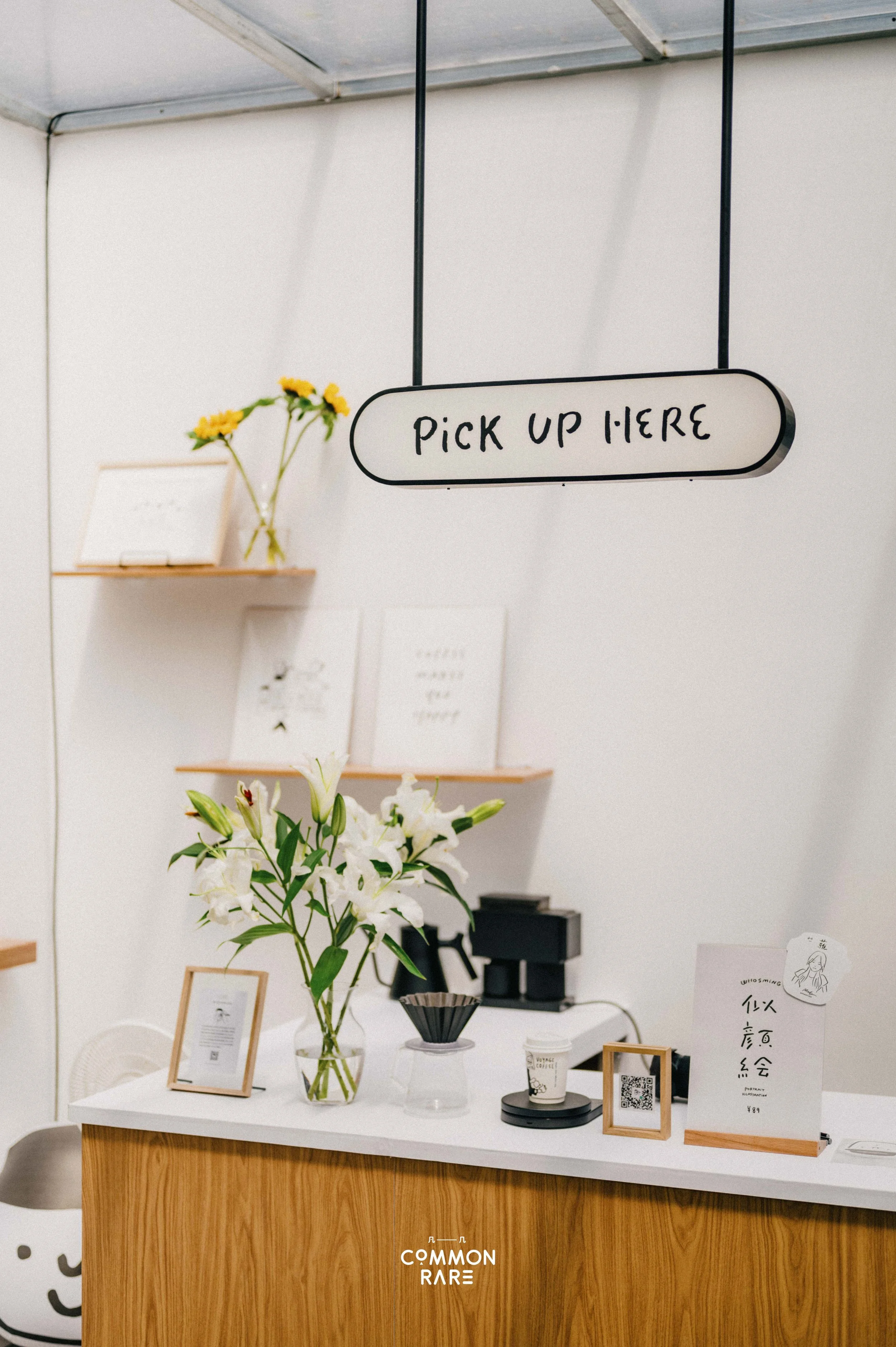 A checkout counter with flowers, framed artwork, paper cups, and signs, located in a minimalist store or cafe setting.