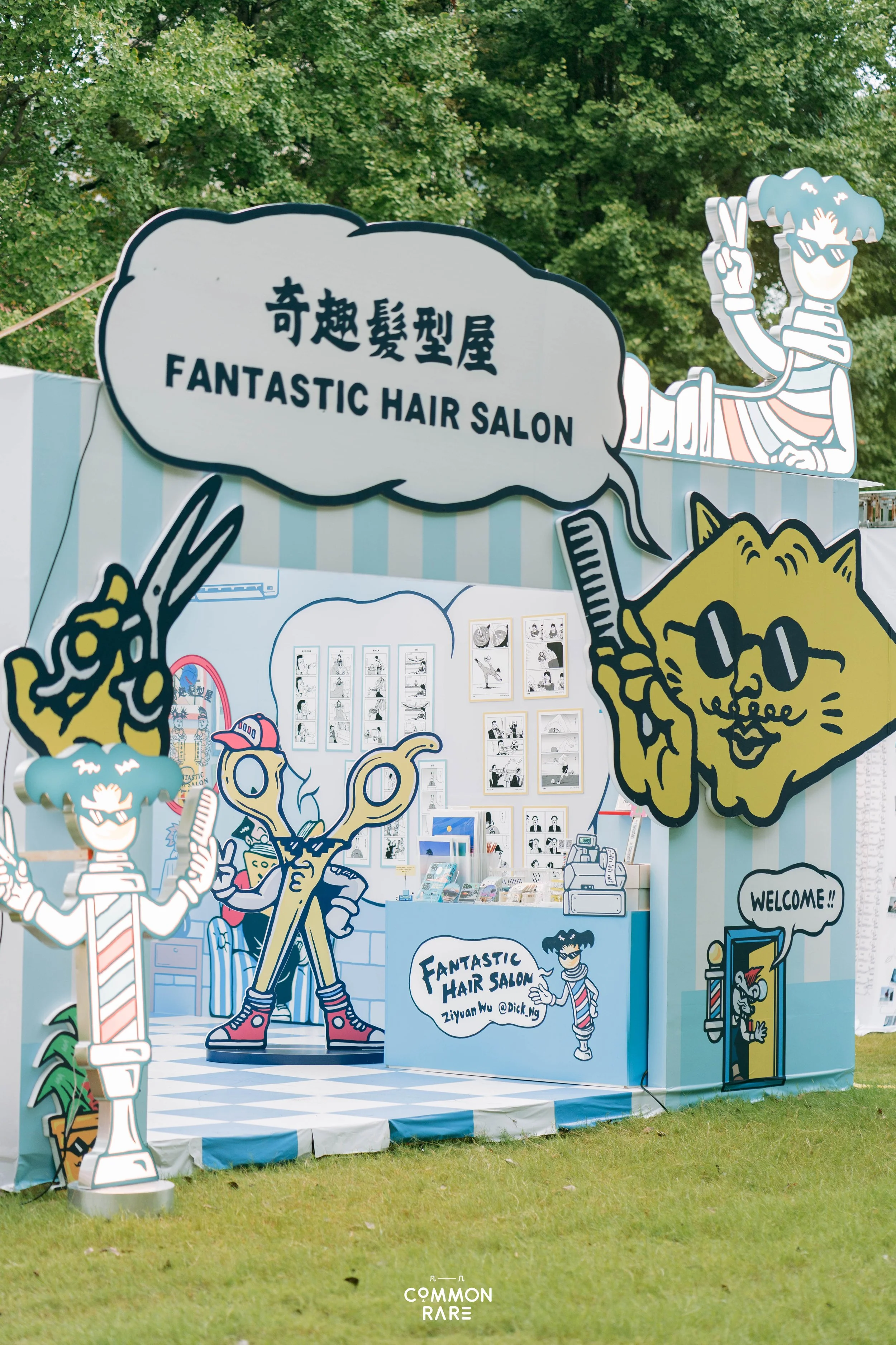 Colorful outdoor booth with cartoon-style cutouts advertising a hair salon called 'Fantastic Hair Salon.' The booth features playful illustrations of characters with exaggerated hairstyles, sunglasses, and various accessories, set against a backgroun
