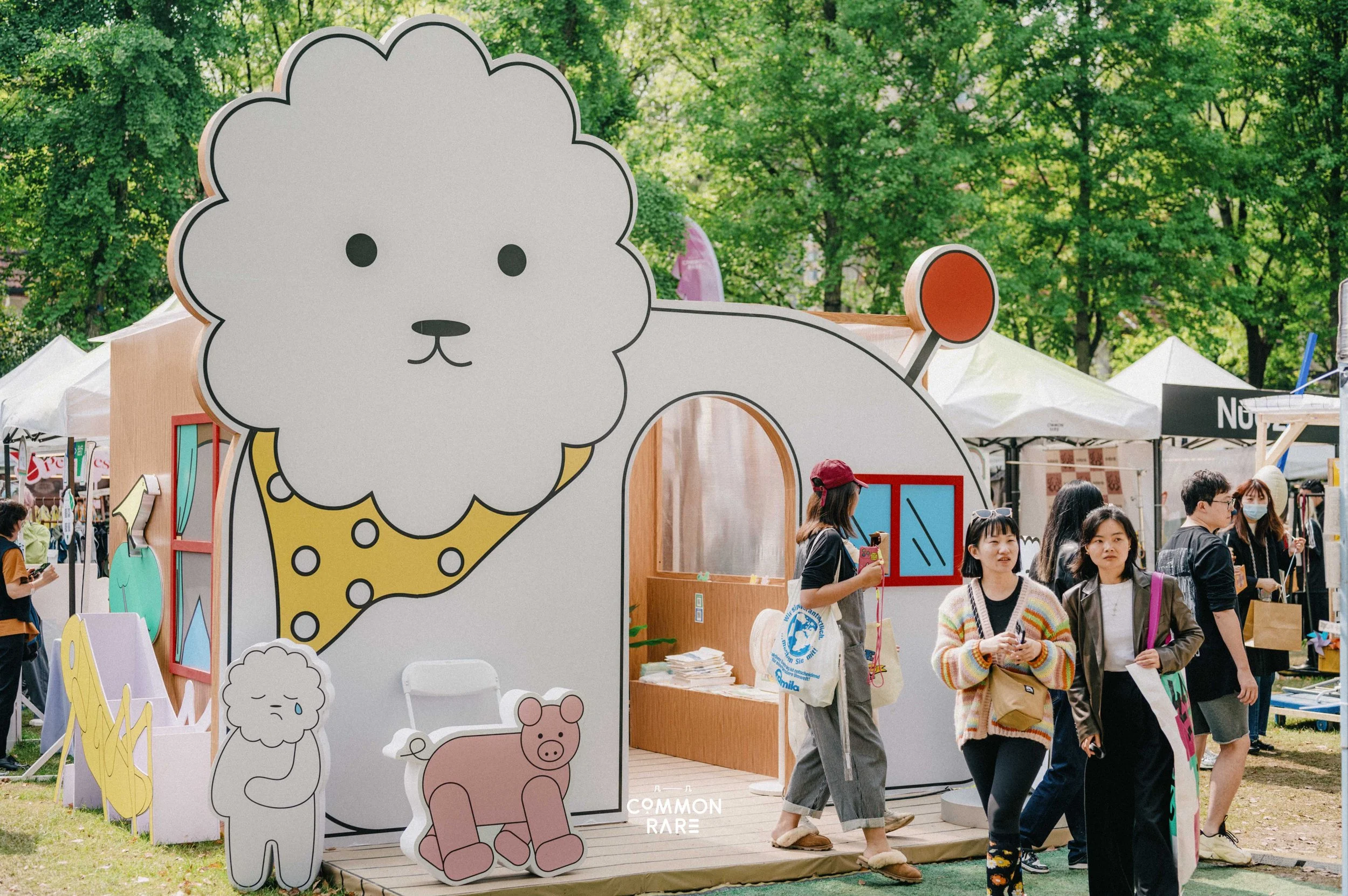 A large cartoon-style display featuring a white fluffy dog with a happy expression, a pink pig, and other small cartoon animals, at an outdoor event with tents and people walking by.