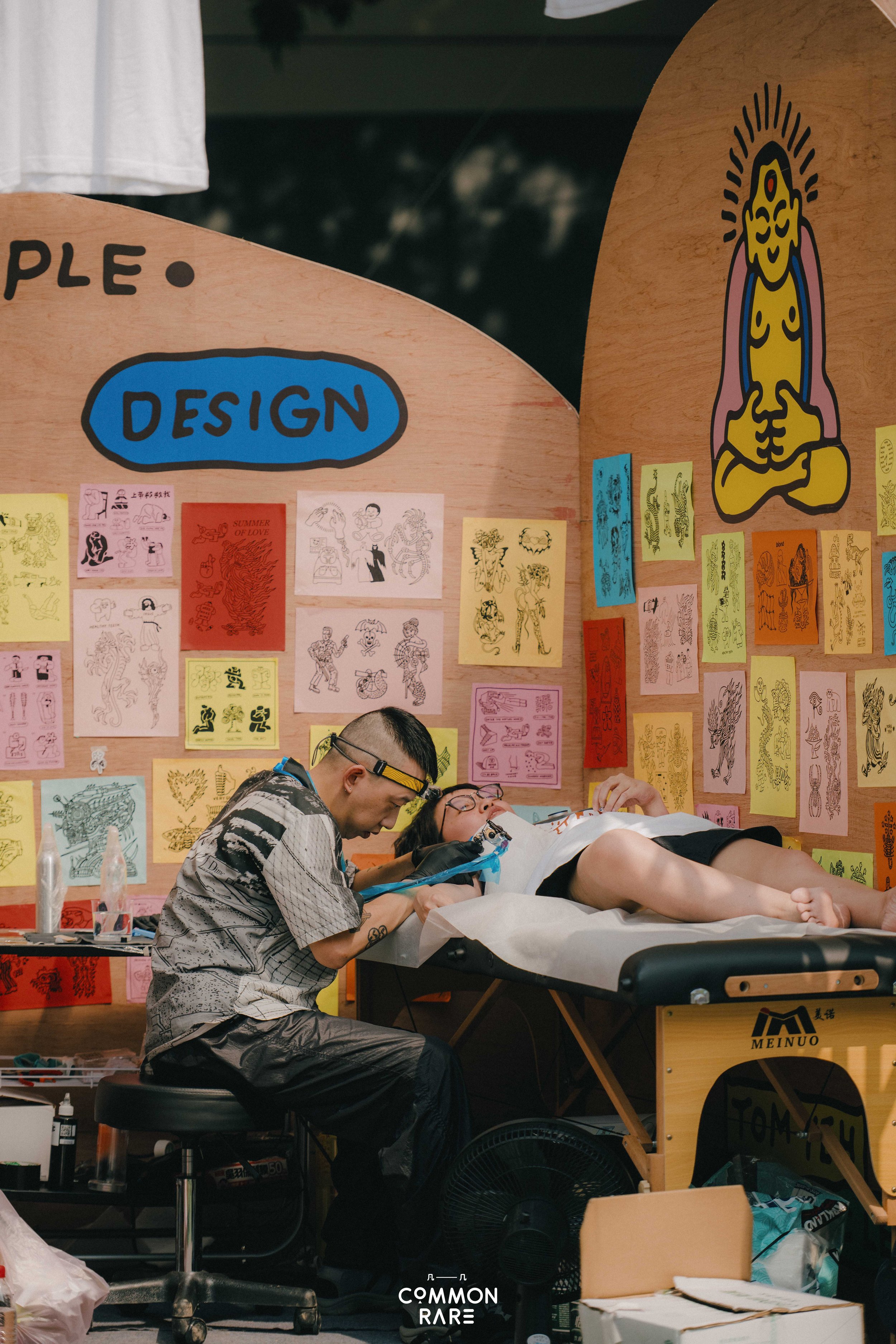 A tattoo artist is tattooing a person lying on a tattoo bed at a tattoo convention. The background features colorful tattoo flash art on the walls and a large yellow poster reminiscent of traditional tattoo styles.
