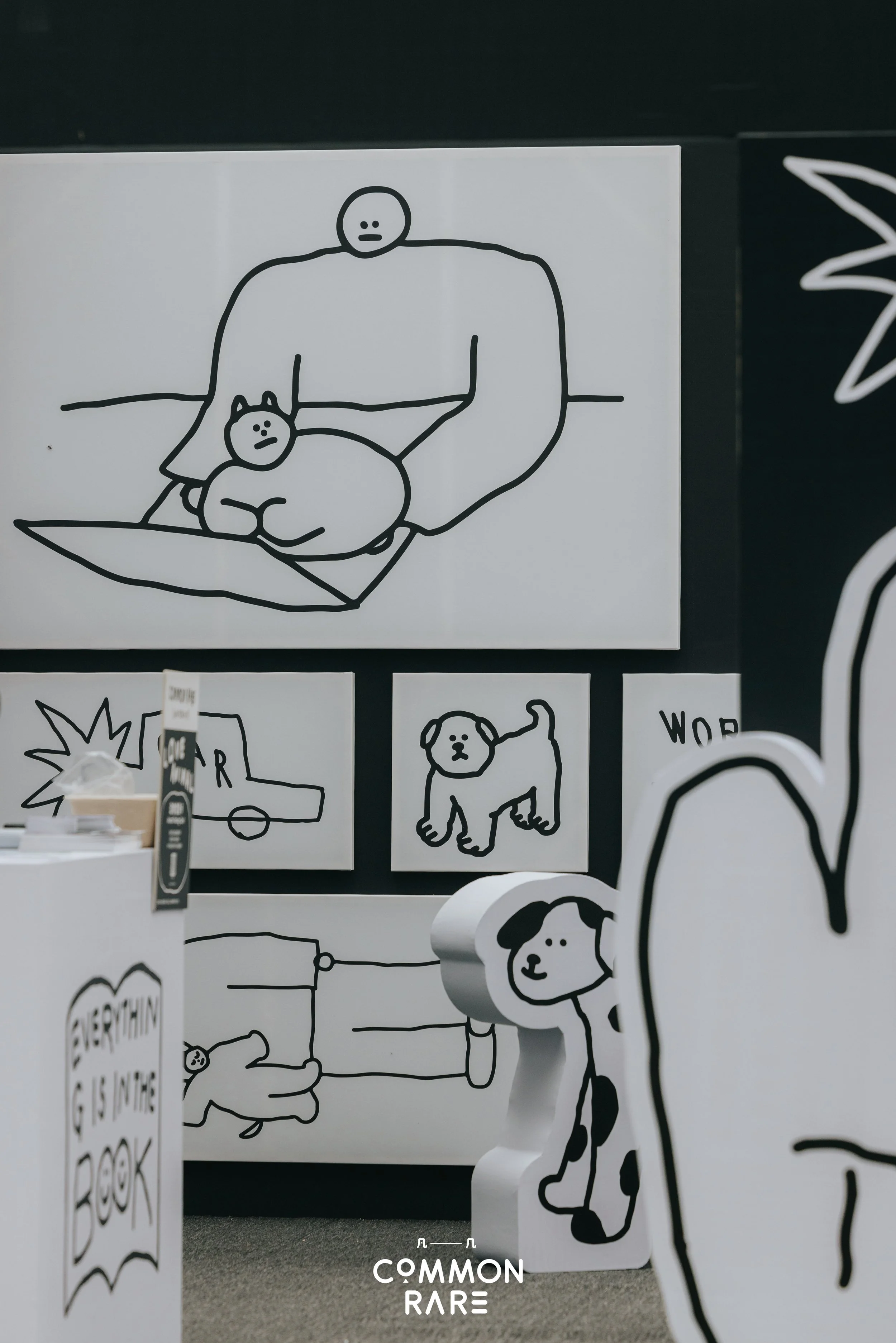 Black and white line drawing of a person sitting on a cushion with a dog on their lap, both with surprised facial expressions. Additional sketches of dogs and text are visible on other artworks in the background.