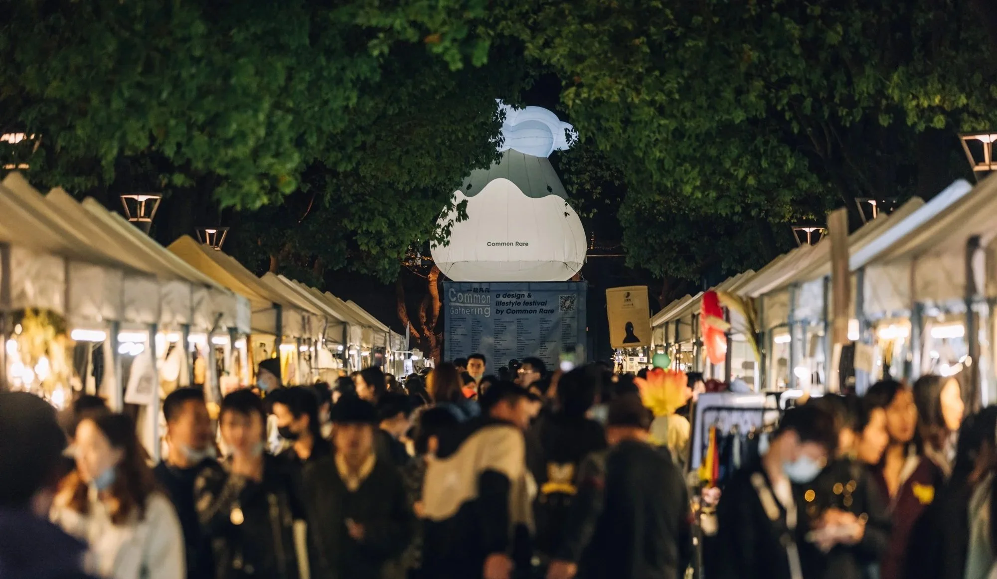 Nighttime outdoor market with crowded shoppers walking between stalls, illuminated by small lights, and a large paper lantern hanging ominously in the background among overhanging trees.