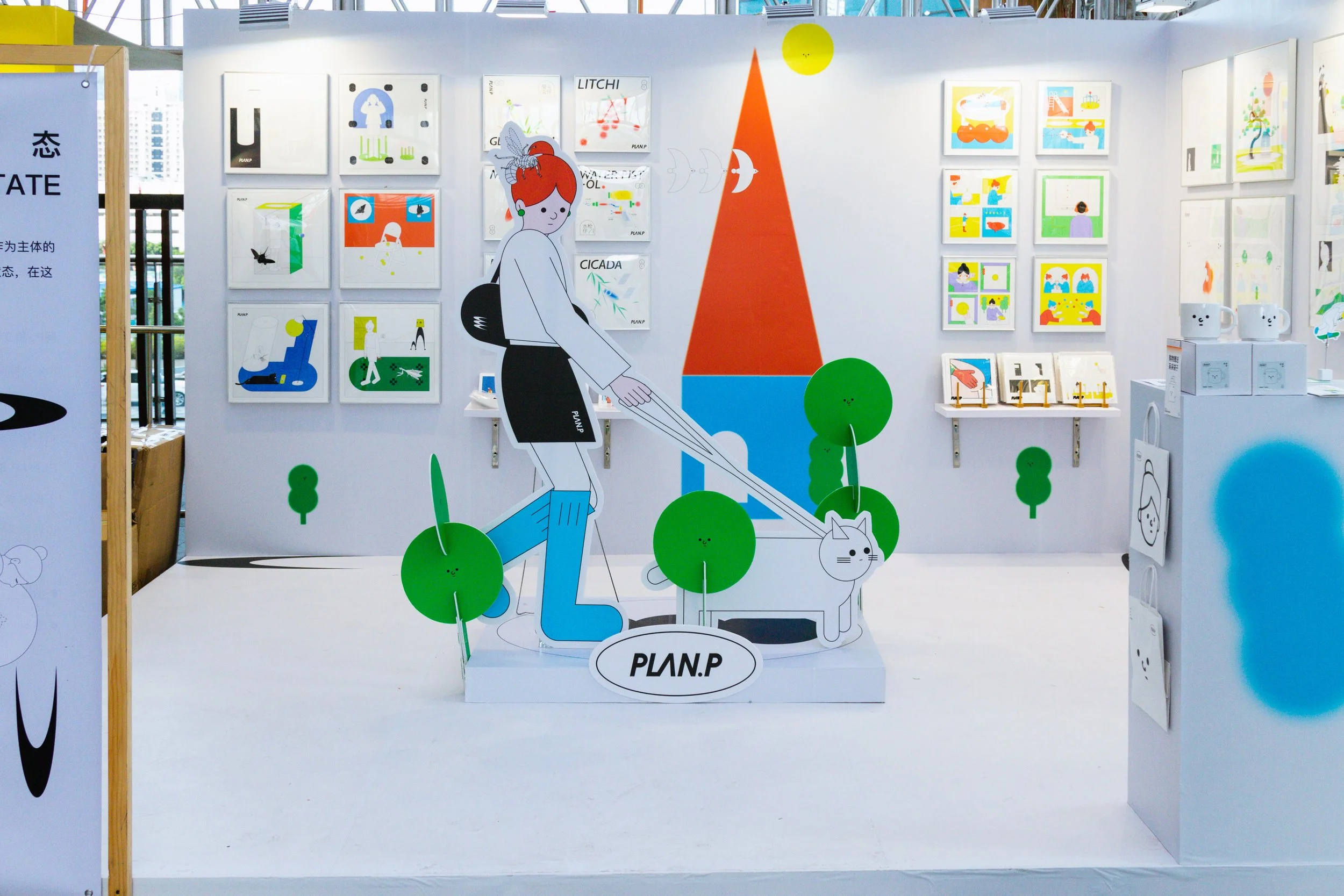 An art display featuring colorful illustrations of animals, people, and abstract shapes on a white wall. There are framed artworks, a large illustration of a person with a bee on their head, and several small artworks and products on tables to the si