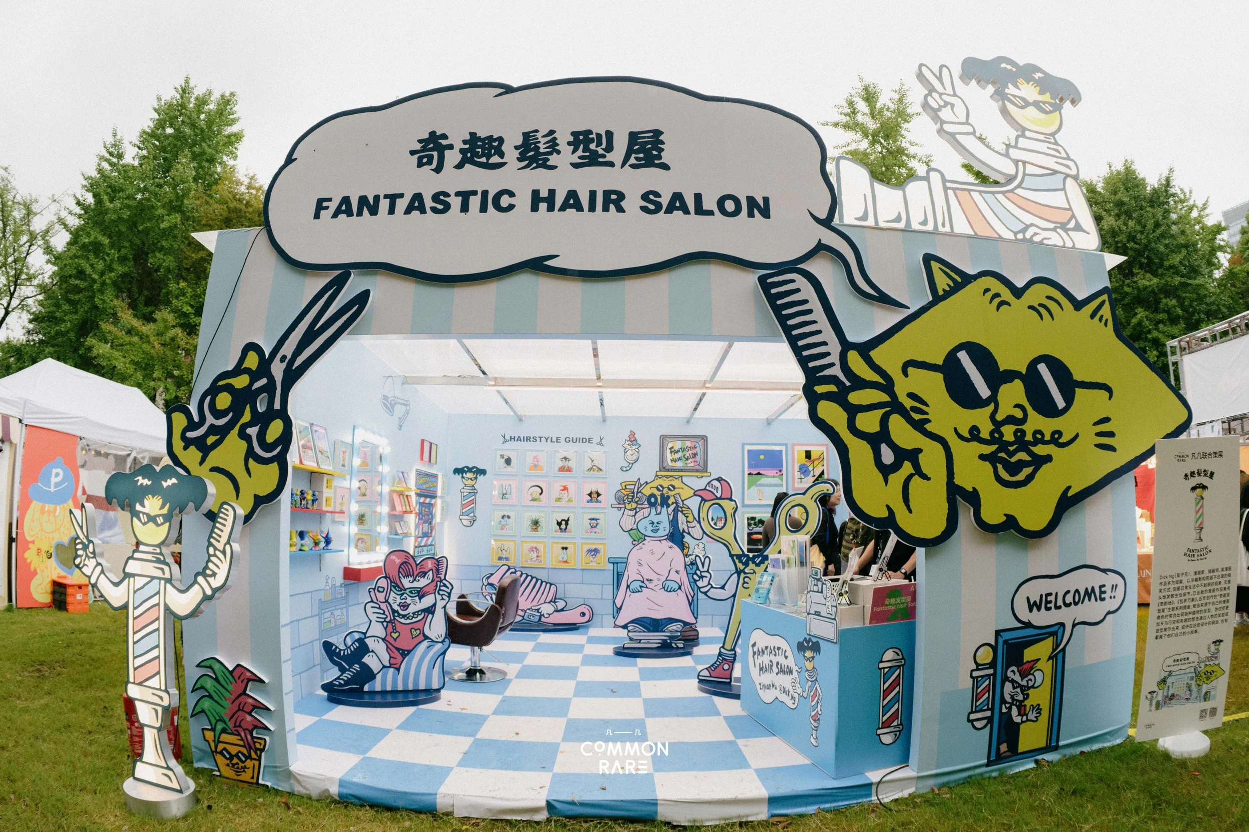 Colorful outdoor booth with cartoon-style illustrations promoting a hair salon, featuring characters with exaggerated hairstyles and accessories, and a sign that reads 'FANTASTIC HAIR SALON.' There are trees in the background, and the booth has a wel