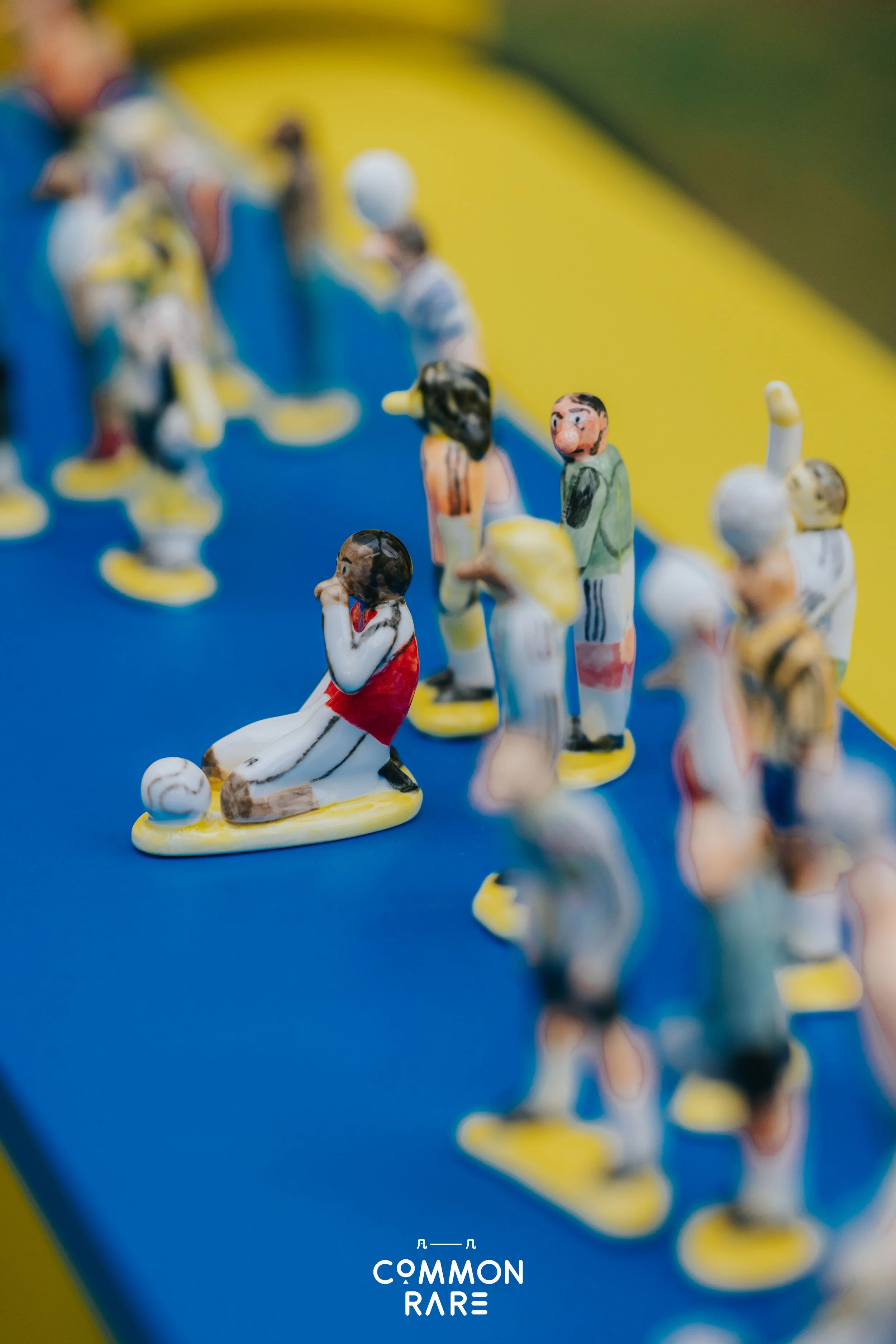 Close-up shot of small, colorful figurines representing soccer players on a blue surface with a yellow background. The figurines are arranged in a line, with one player kneeling and others standing, some holding hands or gesturing. The text 
