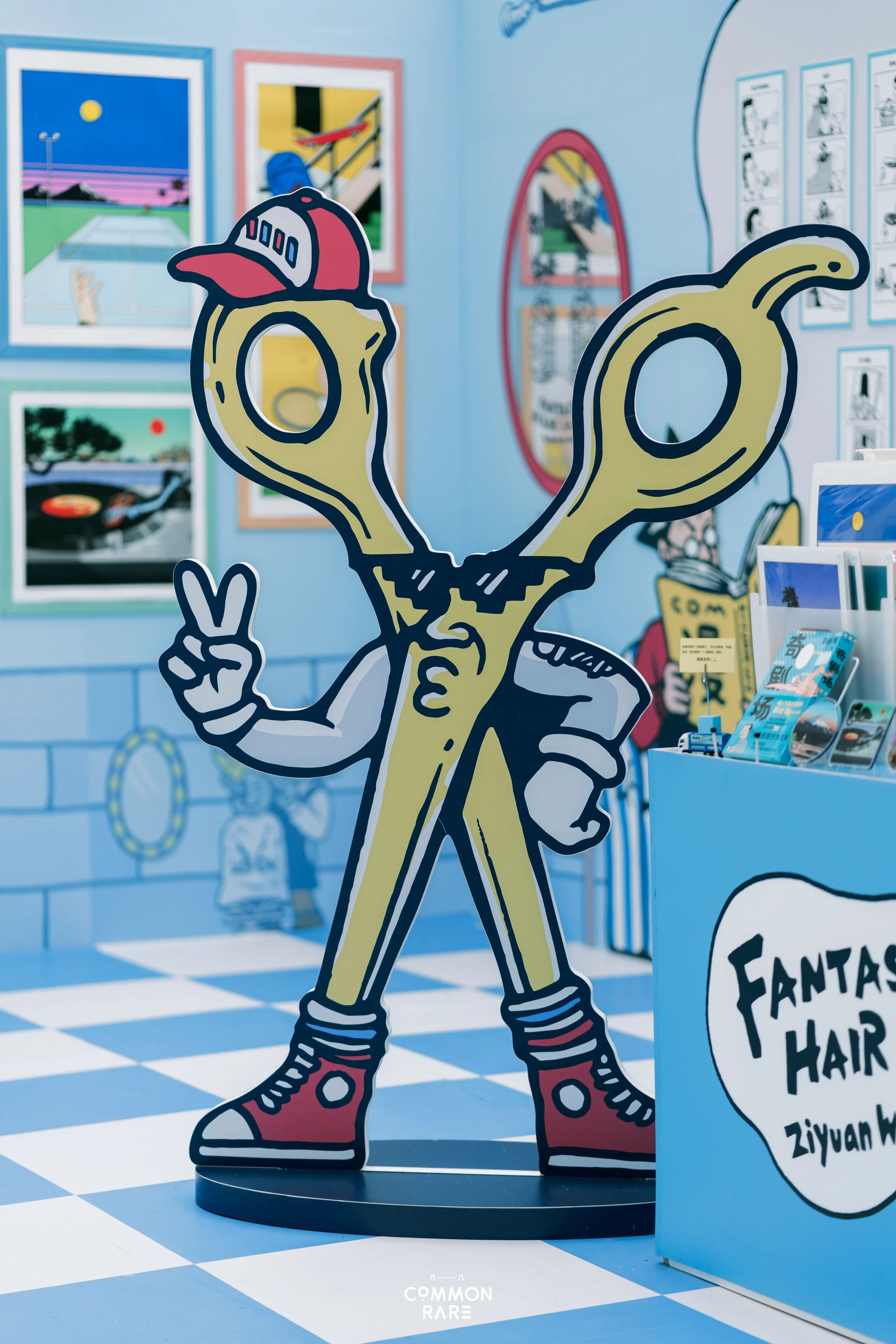 Colorful cartoon-style cutout of a character with large eyes, wearing a cap, making a peace sign, in a vibrant art gallery or exhibition space with framed artwork on the walls.