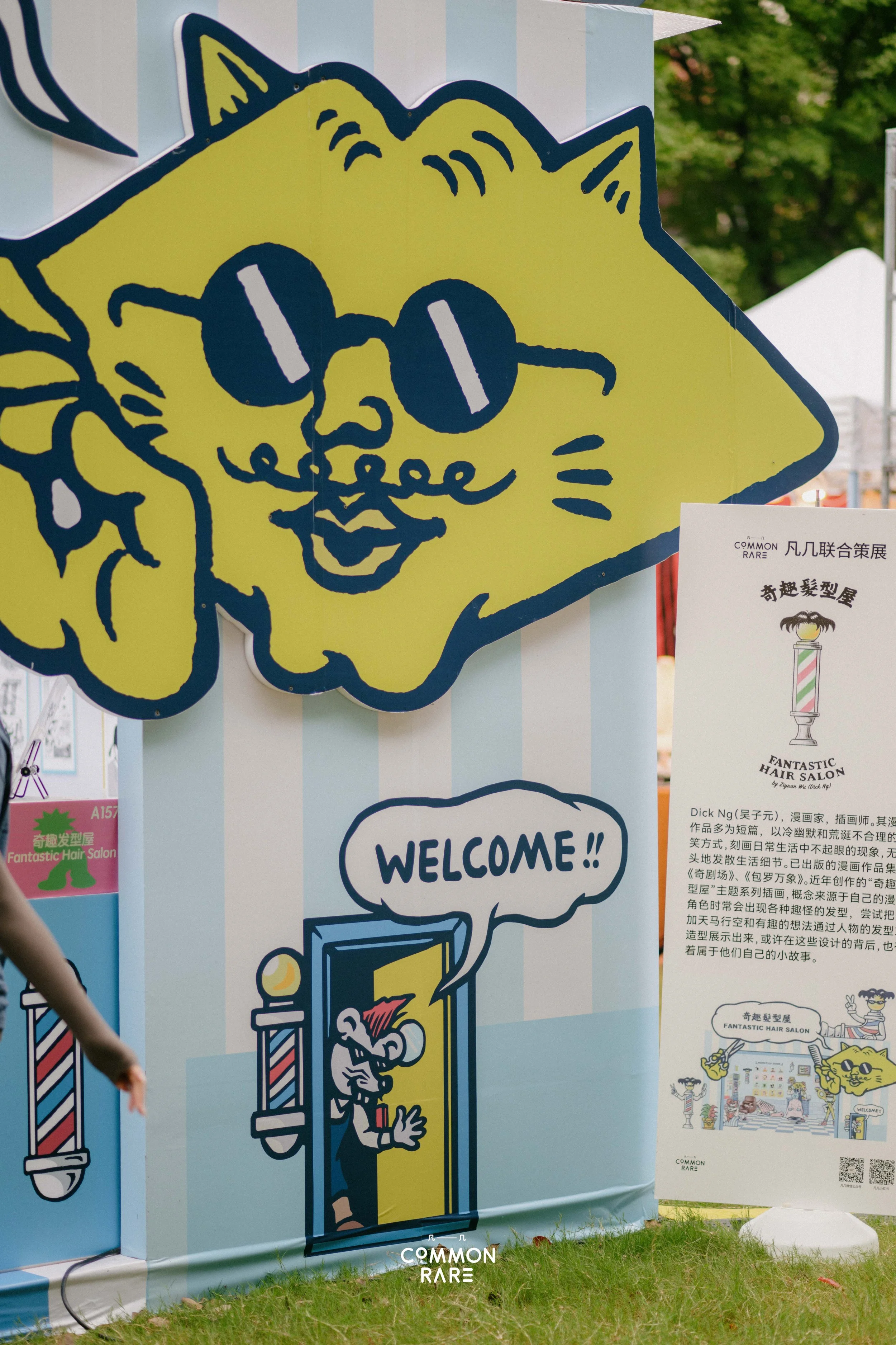 Colorful cartoon-style sign with a yellow cartoon cat wearing sunglasses, sitting behind a barber pole topped with a pineapple, and a speech bubble saying "WELCOME!!" in bold letters. The sign is at an outdoor event with trees in the background.