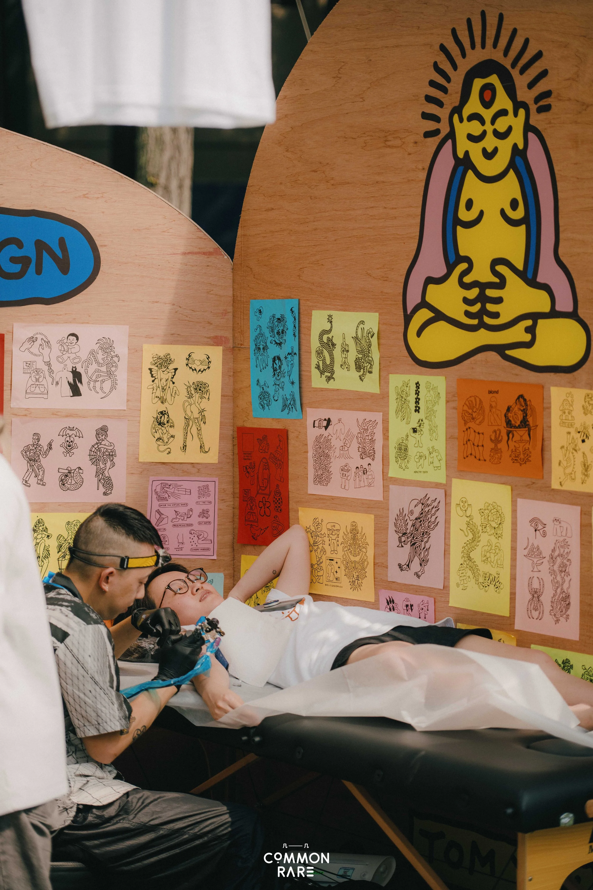 Tattoo artists are tattooing a person lying on a table in front of a wall decorated with colorful drawings of various tattoos. One artist is working on the person's leg, while the other is observing or preparing. The wall features a large illustratio
