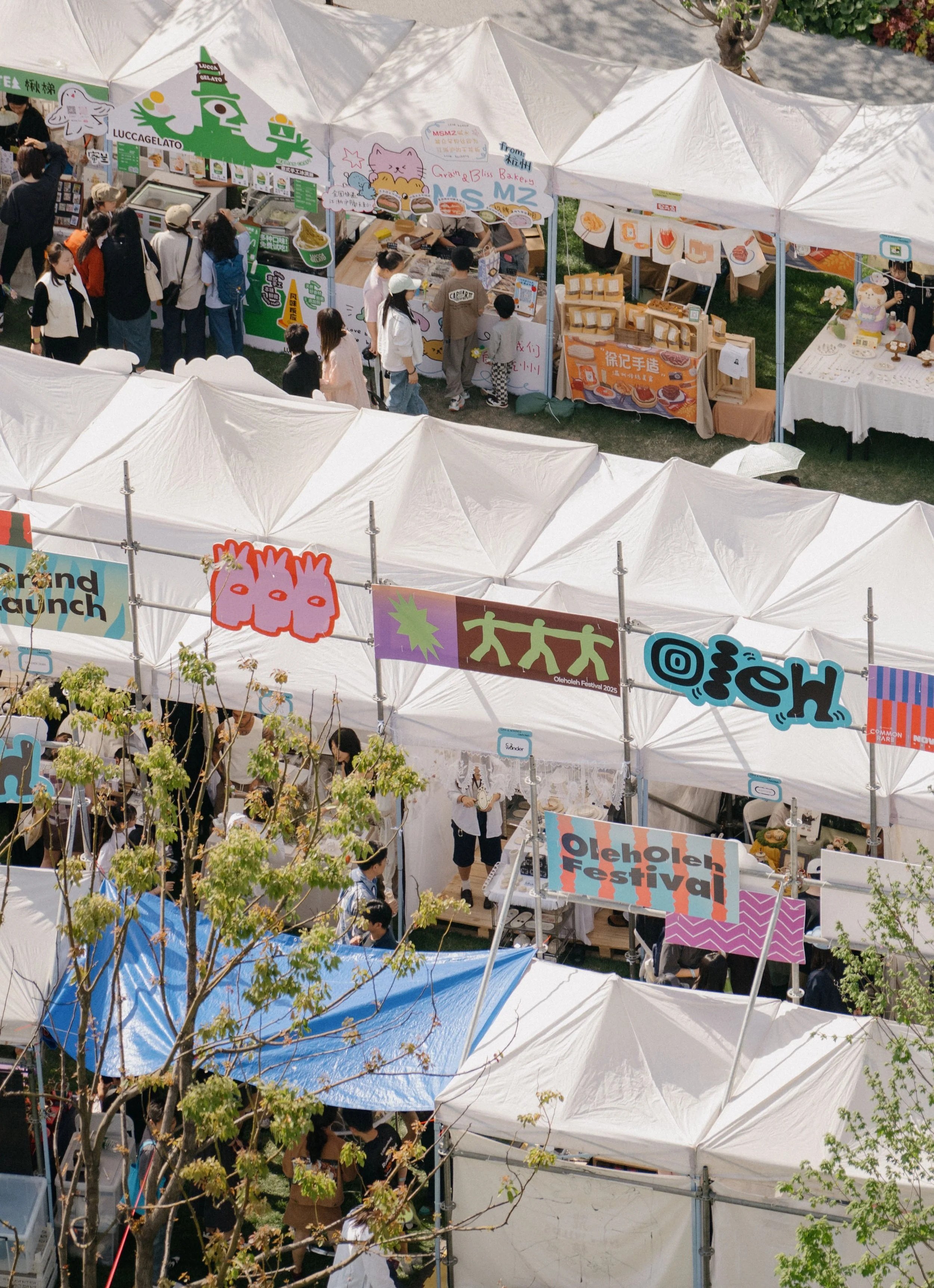 Overhead view of a bustling outdoor festival with white tents and colorful banners, and a crowd of people browsing various stalls.