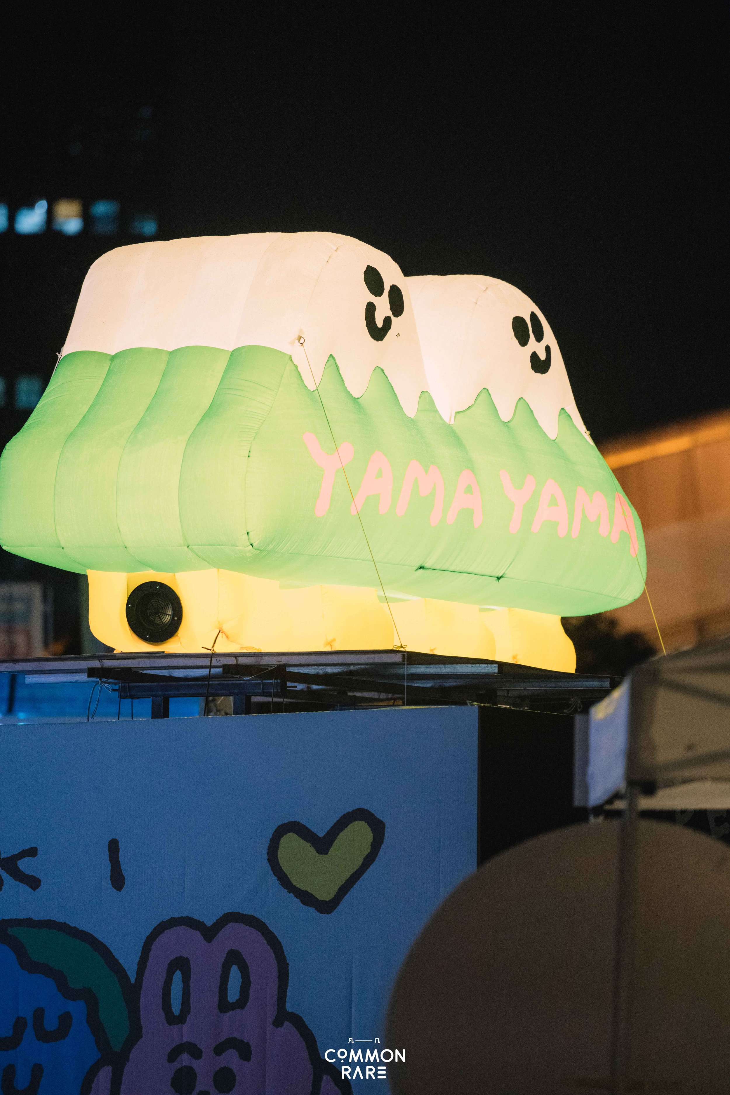 A sky lantern illuminated from within, decorated with a stylized ghost face and the words 'YAMA YAMA' in pink text, floating at night against a dark sky background.