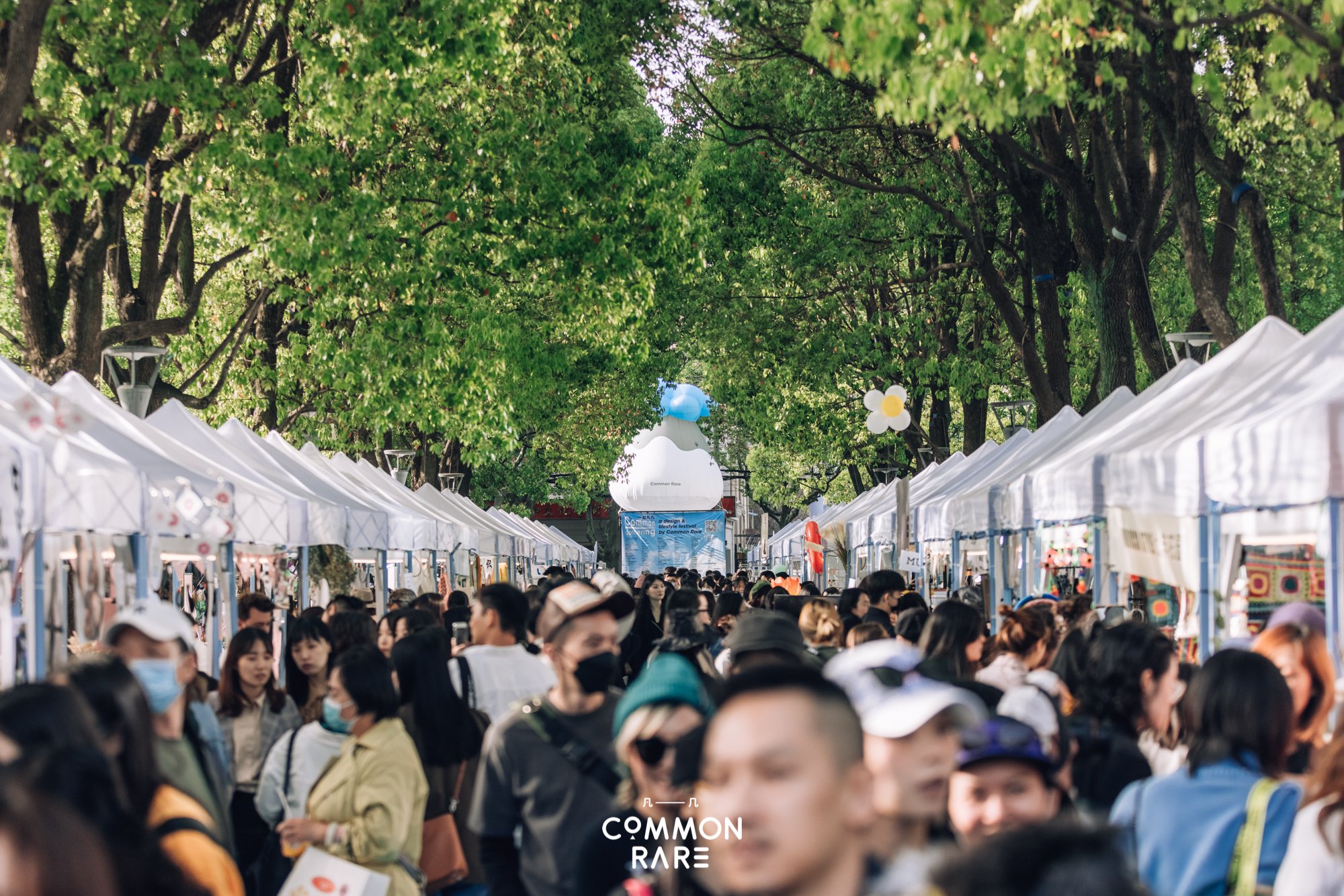 Crowd of people walking along outdoors market stalls with white tents, lined with green trees overhead, at a festival or event.