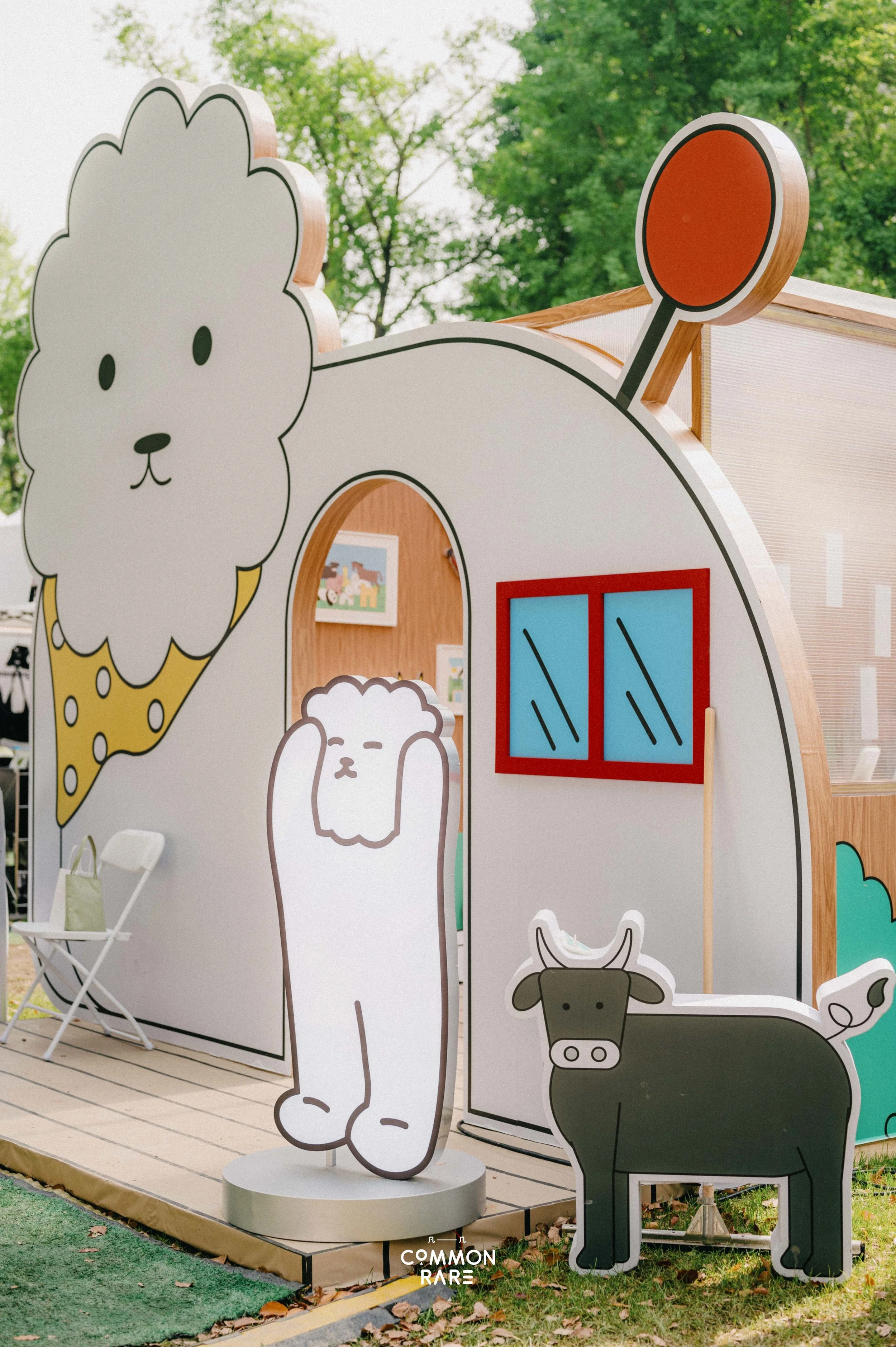 Colorful cartoon farm animal cutouts including a sheep, a cow, and a pig, displayed outdoors on a wooden platform with a small seating area nearby. Trees in the background.
