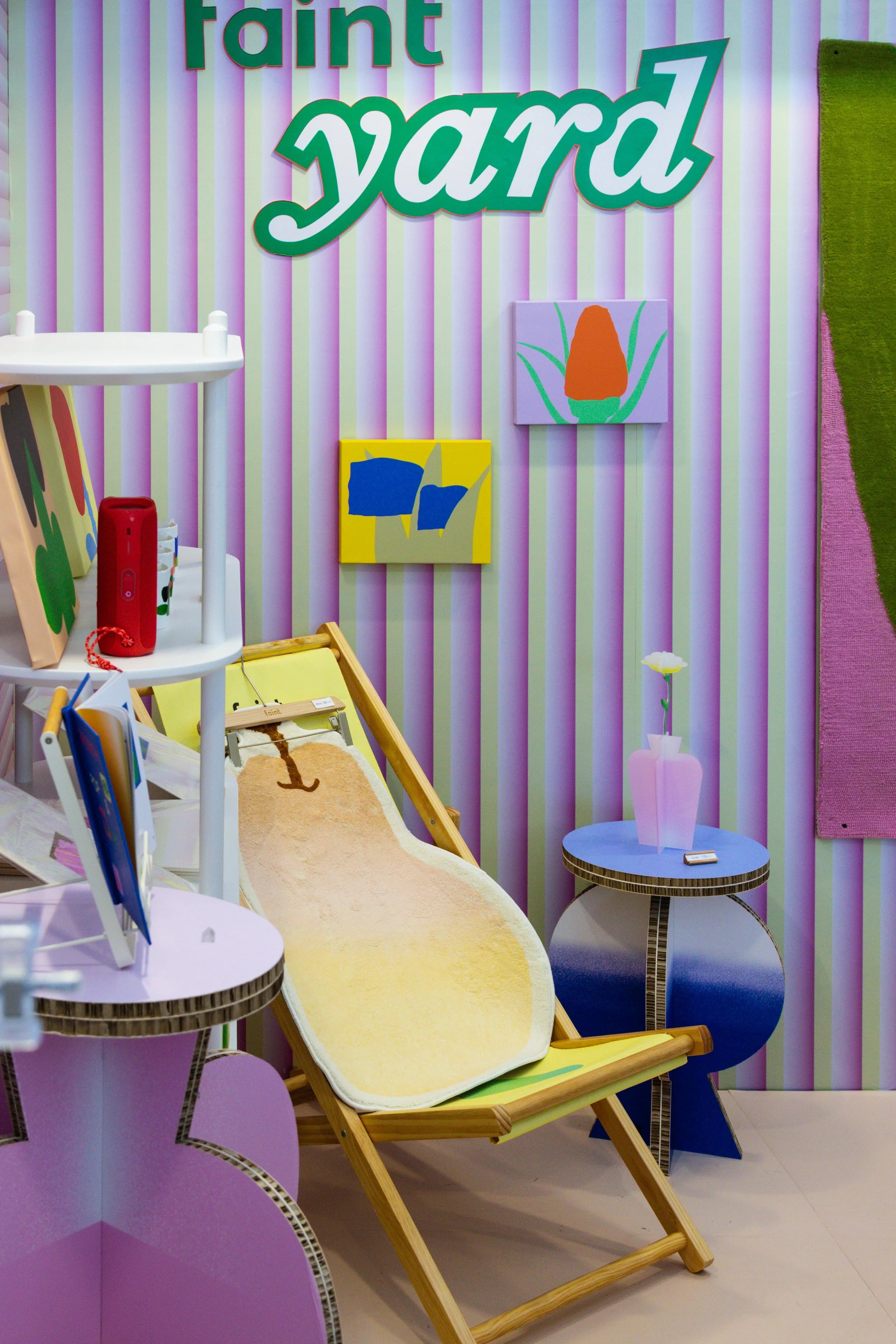 Colorful display with a striped pink and white wall, decorated with purple, green, orange, and blue artwork and signs with the word 'faint yard.' There is a white shelf with books and a red speaker, and a beige lounge chair shaped like a pear with an