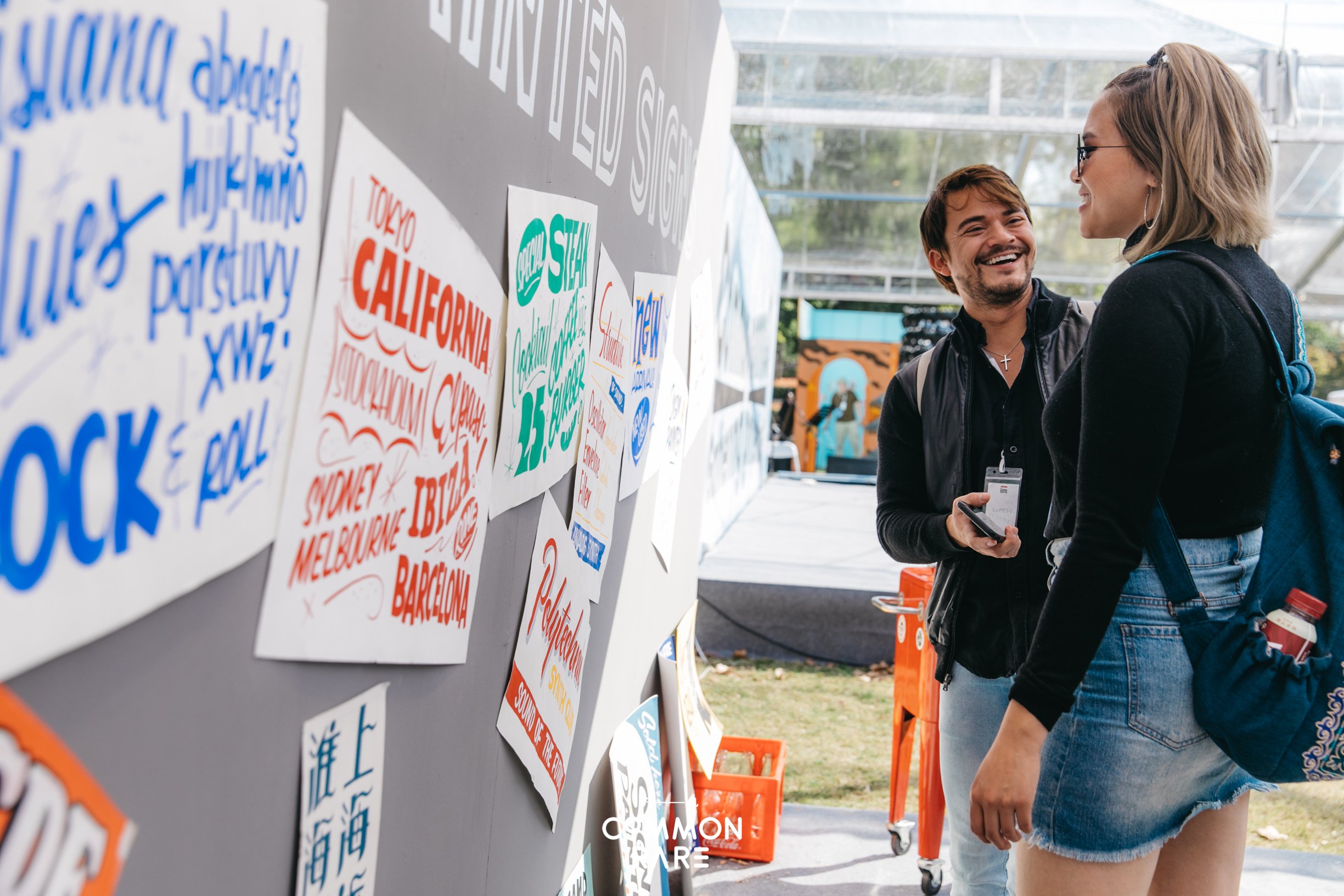 Two people, a man and a woman, are engaged in conversation at an outdoor event, with colorful signs and written words on display behind them.