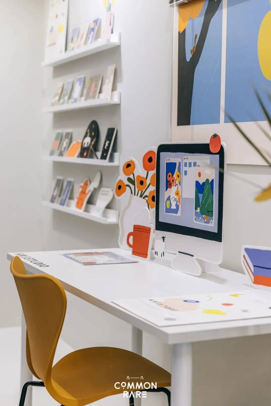 A display setup with a yellow chair in front of a white table, featuring a computer and decorative paper cutouts of flowers and a jar. In the background, there are white shelves with books or magazines and wall art with abstract and nature themes.