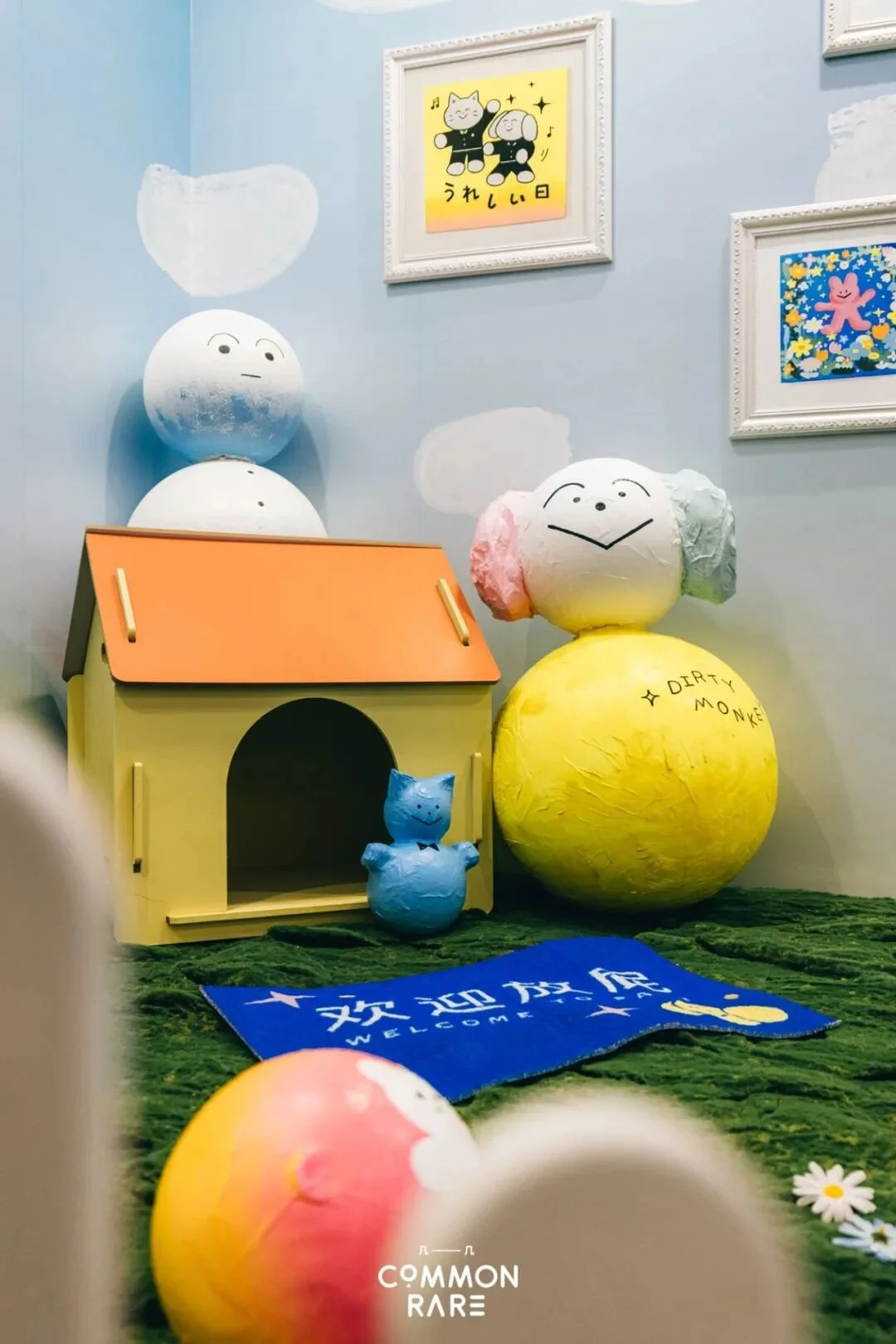 Colorful display of cartoon character figures, including one with colorful puffy hair, a blue cat, and a face with a big smile, in a room with framed artwork on the walls and a blue welcome mat on the floor.