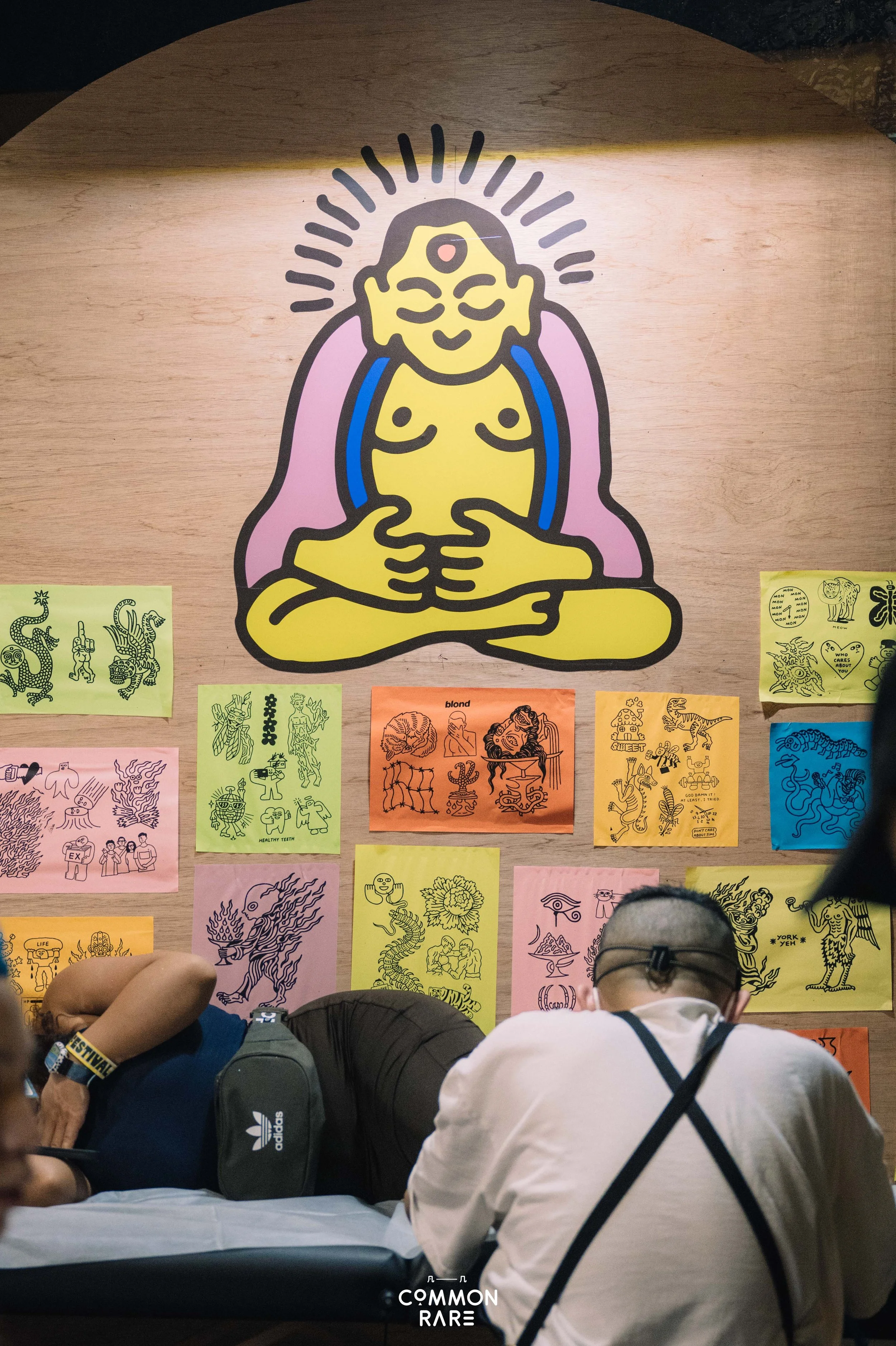 People sitting at tables in front of a wall with colorful posters of various line art illustrations and Asian-inspired designs, including mythical creatures and characters, in a possibly indoor cafe or gallery setting.