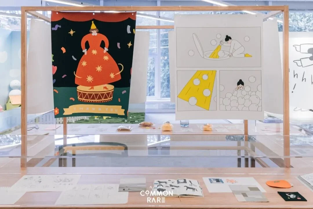 A display with colorful illustrations and crafts, including a large banner of a person in a red dress on a drum, and black and white drawings of a girl with bubbles and cheese, set up in a bright room.