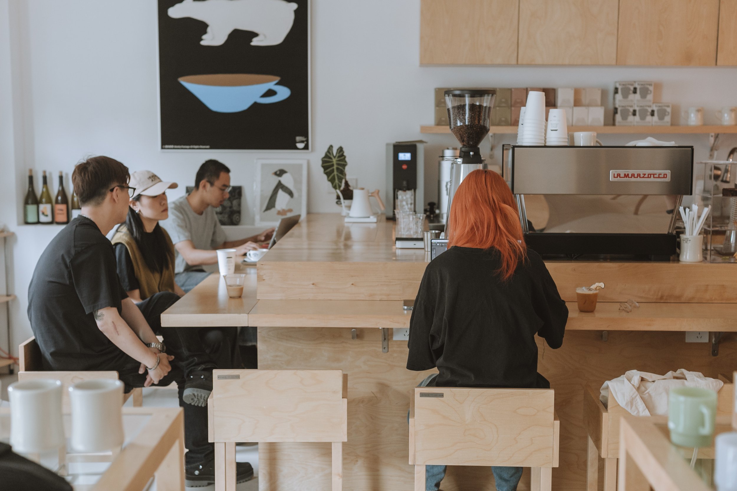 People sitting at a wooden counter in a coffee shop, with a barista behind the counter, espresso machine on the counter, and coffee beans in a grinder.