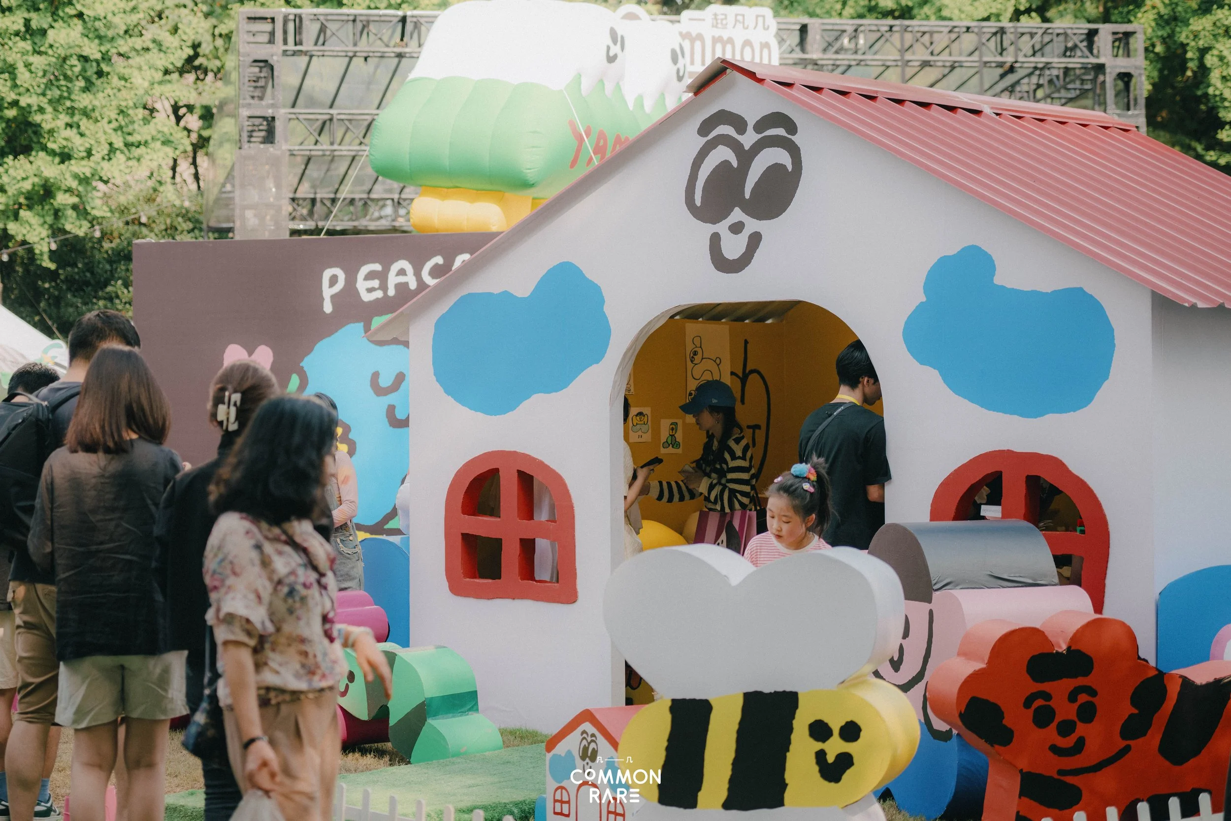 People gather around a colorful toy house with animal illustrations, at an outdoor event with trees and a stage in the background.
