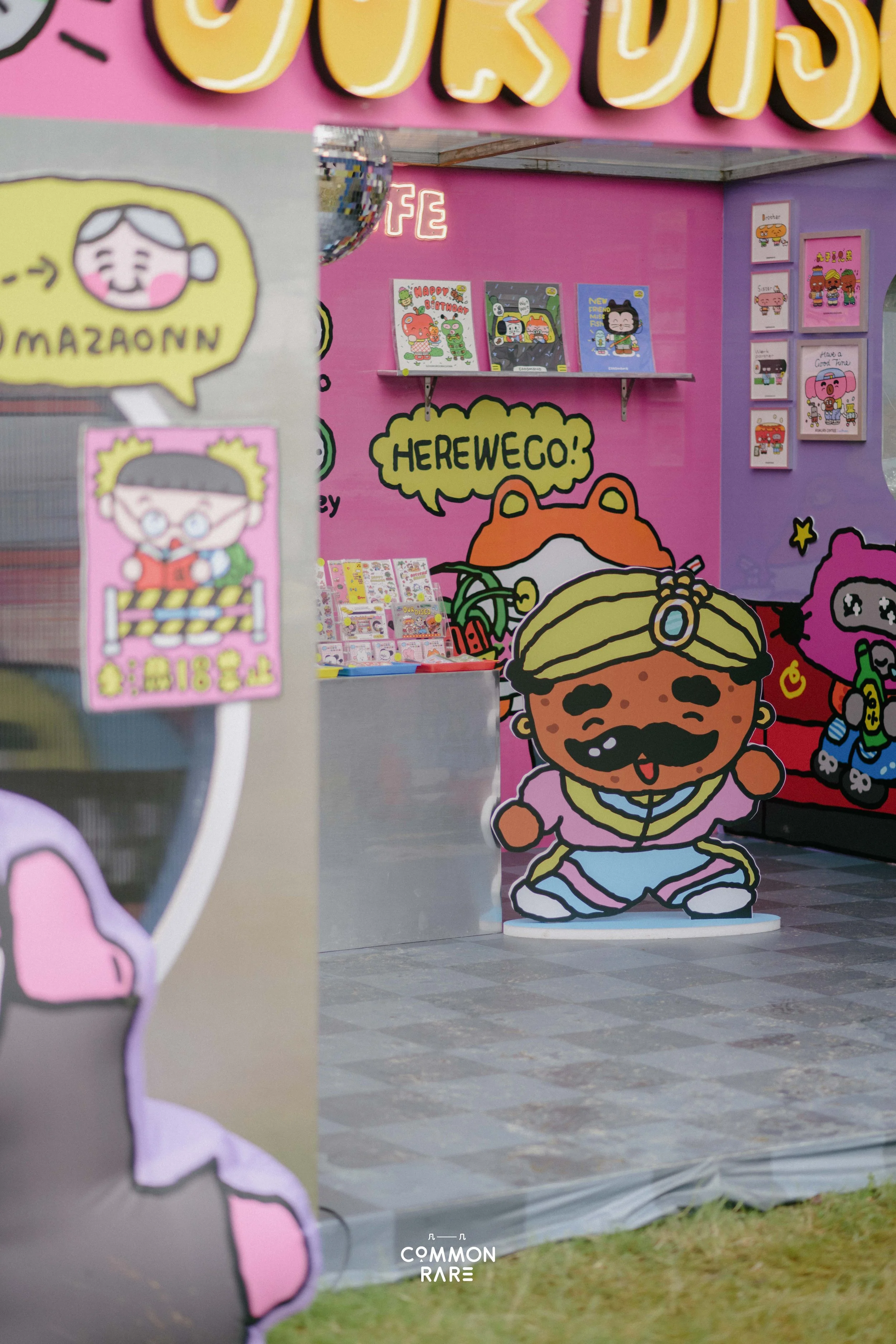 Colorful cartoon characters and illustrations at a booth or display, including a large cutout of a happy cartoon person with a beard, a character with glasses, and various framed images on the wall, with pink, purple, and yellow background colors.