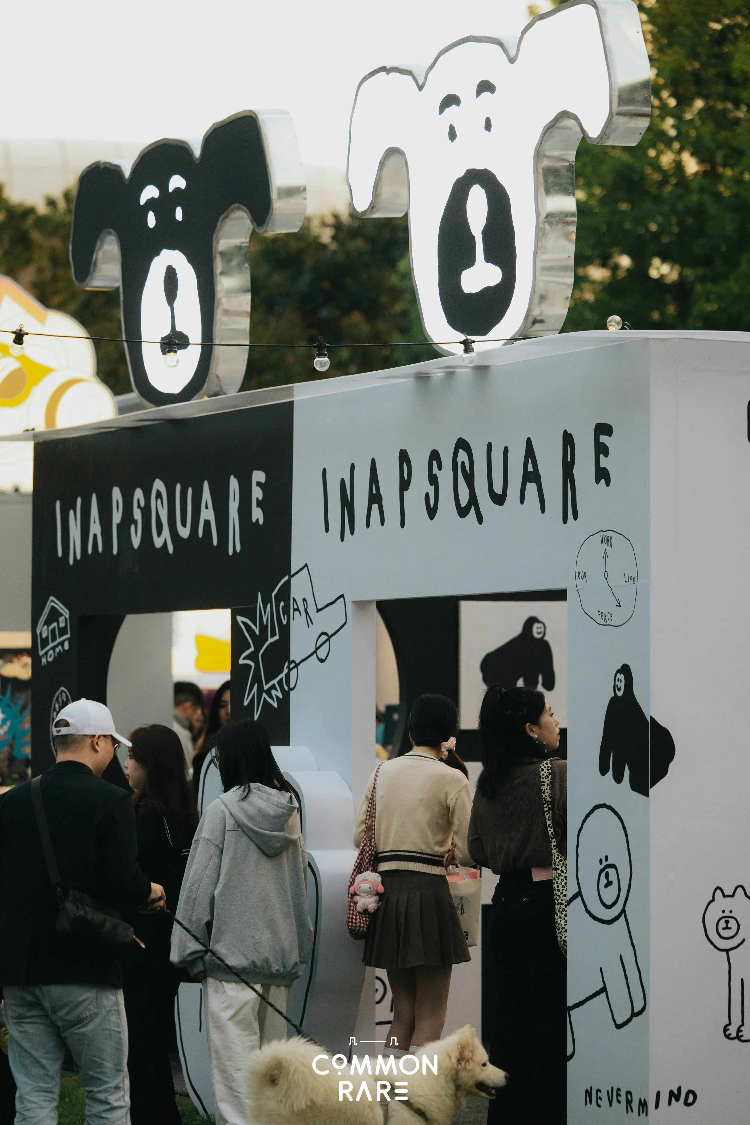 People standing outdoors at a booth with dog-themed artwork and signs, including a large sign reading 'INAPSQUARE' and various dog illustrations, with some individuals holding pets and others observing the display.