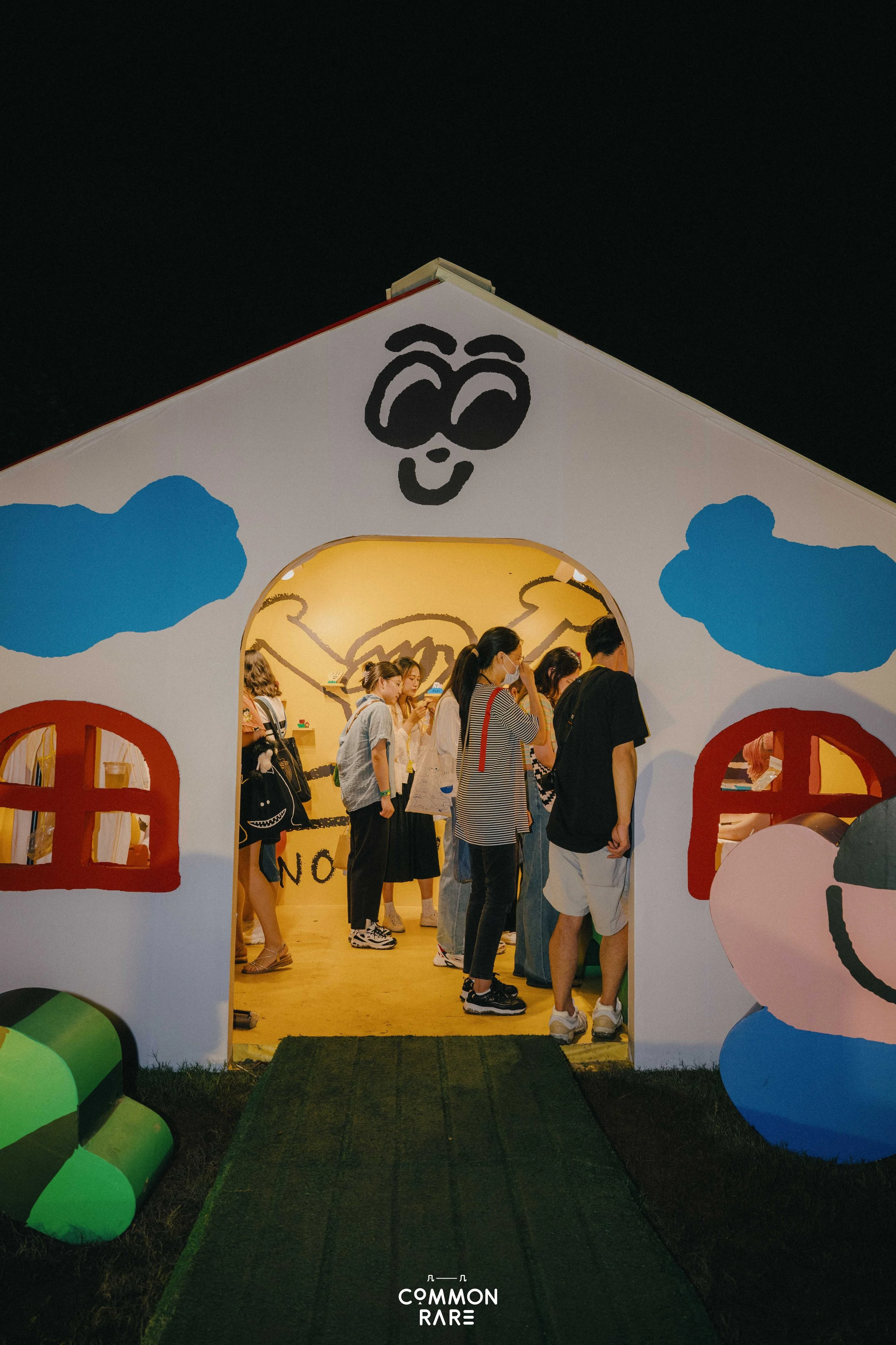 People standing in line inside a whimsical, house-shaped structure with colorful painted decorations, including a cartoon panda face at the top, at night outdoor event.