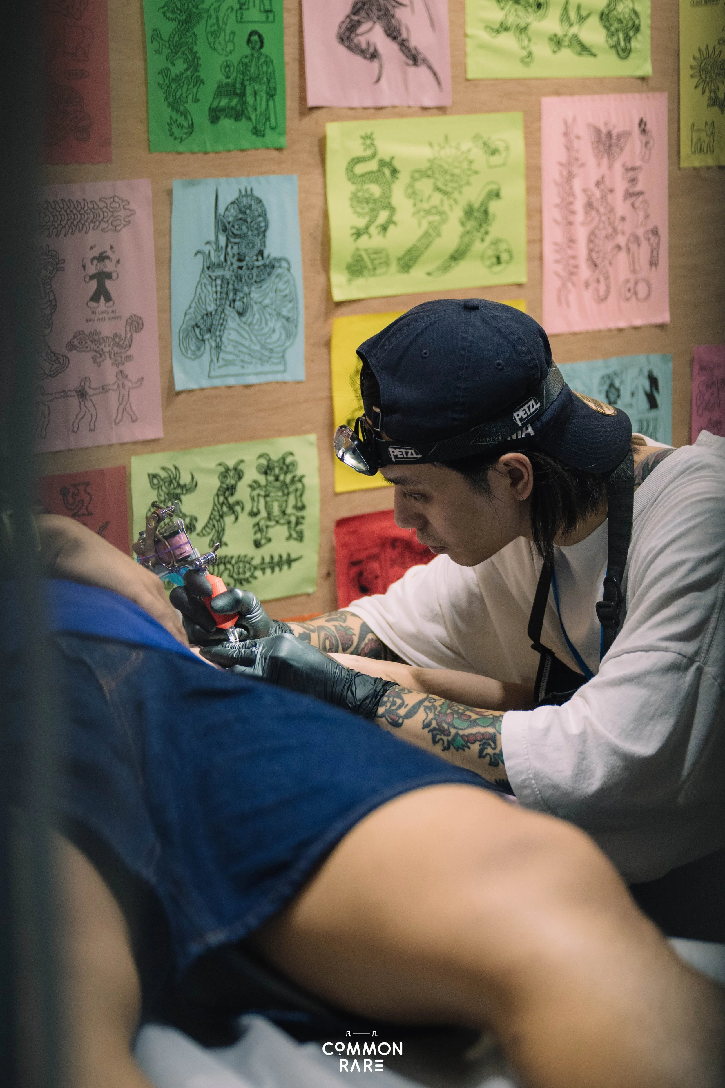 A person tattooing another person's arm in a tattoo shop with colorful tattoo designs on the wall behind them.