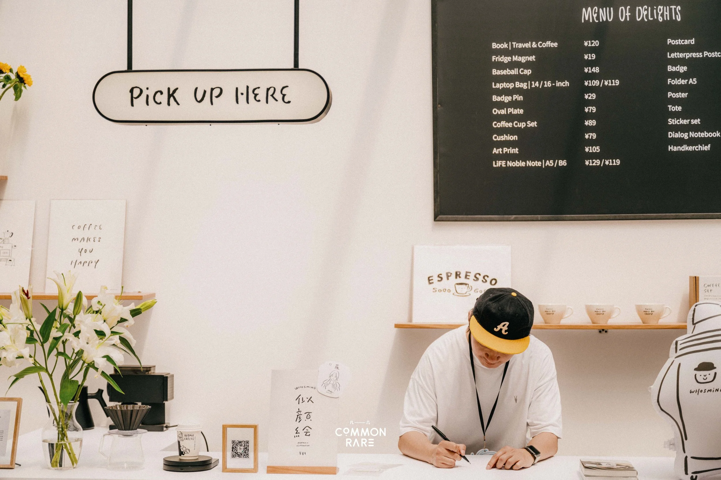 A man wearing a black cap with a yellow brim and a white shirt is writing at a counter in a bright, modern store. There are flowers on the left, a sign reading 'PICK UP HERE' hanging on the wall, and a large menu of items with their prices on a black