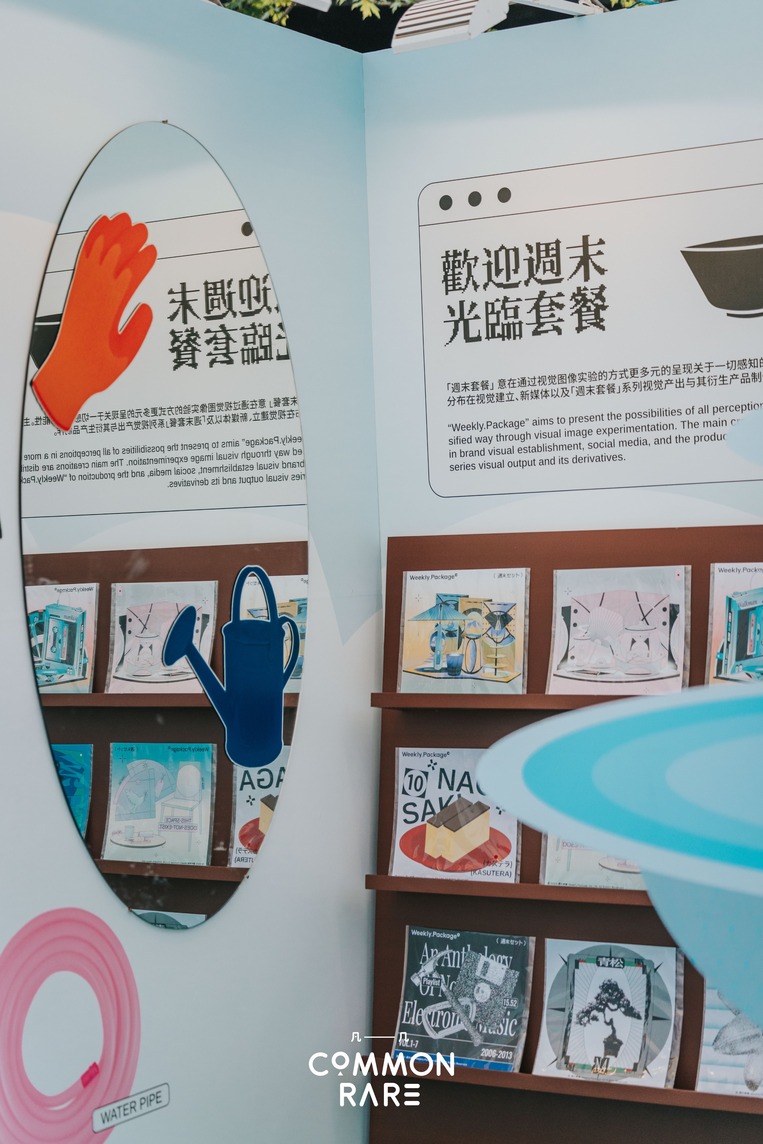 A display with a mirror shaped like a water pipe, a cut-out of a watering can, and several posters about weekly packages and music. The background features a cabinet with books and magazines. The text on the posters is in Asian characters and English