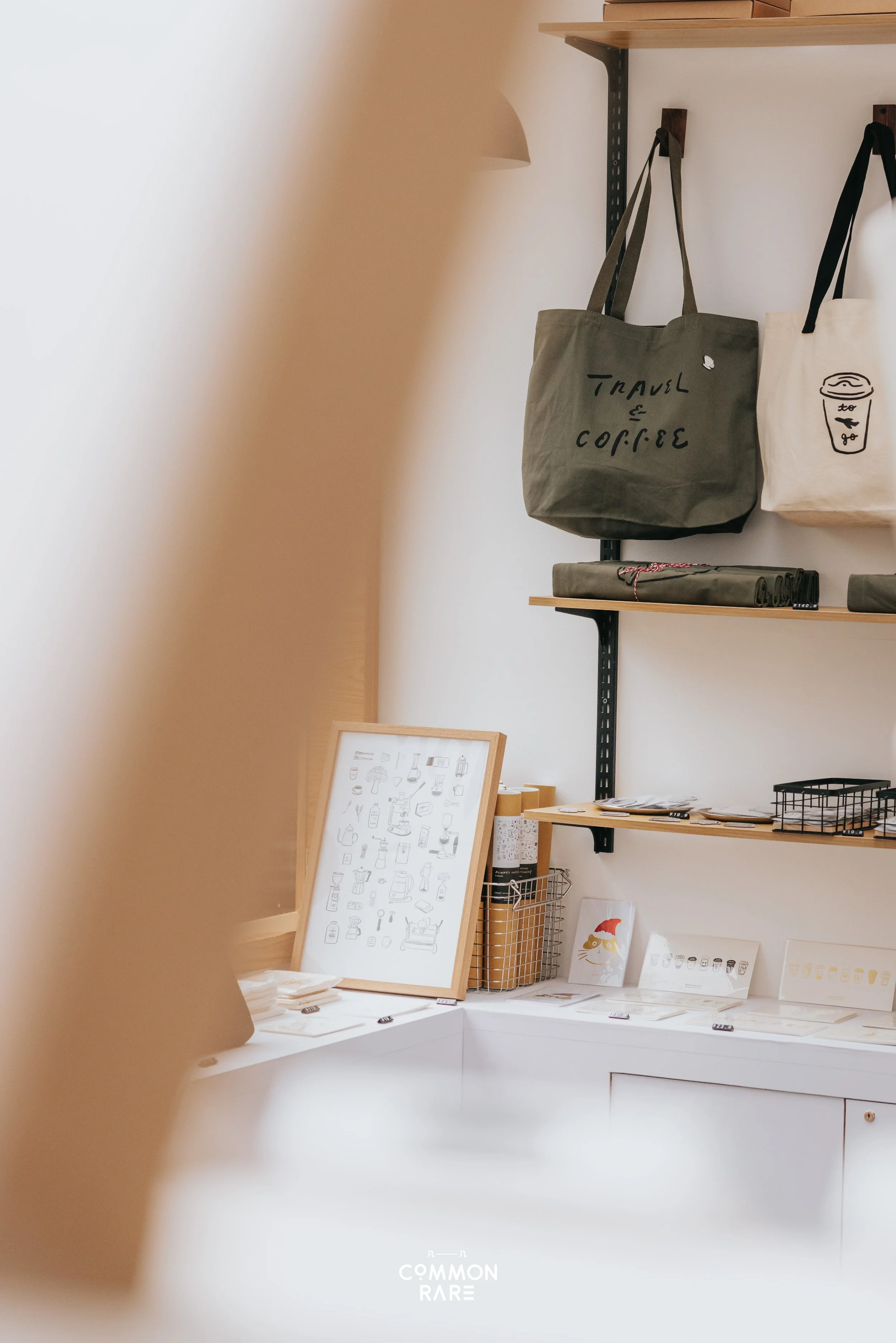 Display of tote bags, a framed art print, and stationery items on a white table in a minimalist store or cafe. The tote bags hang on a black metal and wooden shelf, one with text 'Travel & Coffee' and the other with a coffee cup illustration. The fra
