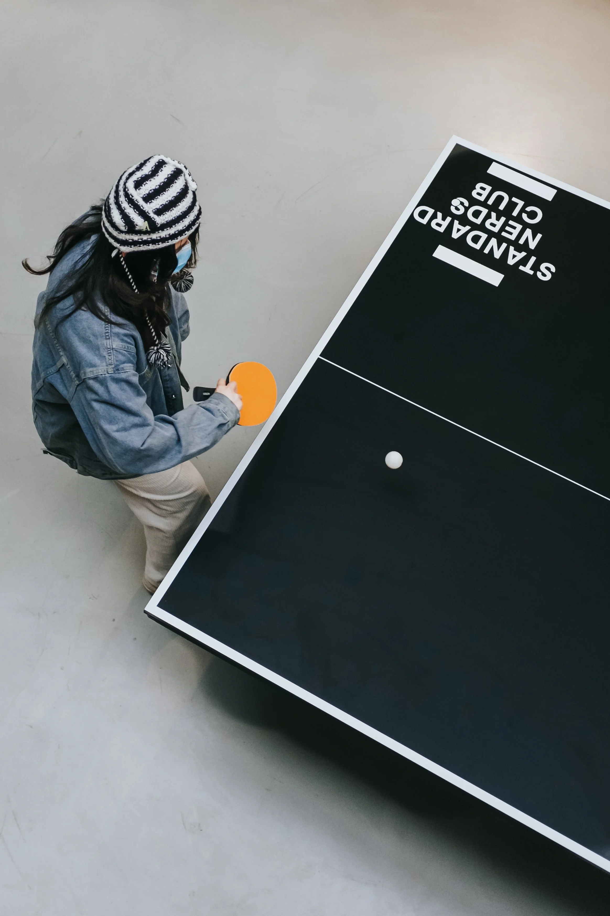 A person wearing a black and white striped beanie, blue jacket, and face mask is playing ping pong on a black table with white edges in a large, open space. The person is holding a yellow paddle and preparing to hit a white ping pong ball.