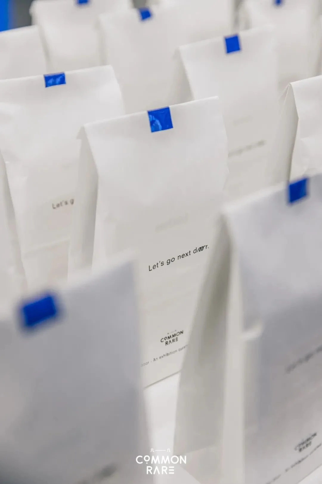 Rows of white paper bags with blue tape closures, some bags have text that says "Let's go next door" and "COMMON RARE."