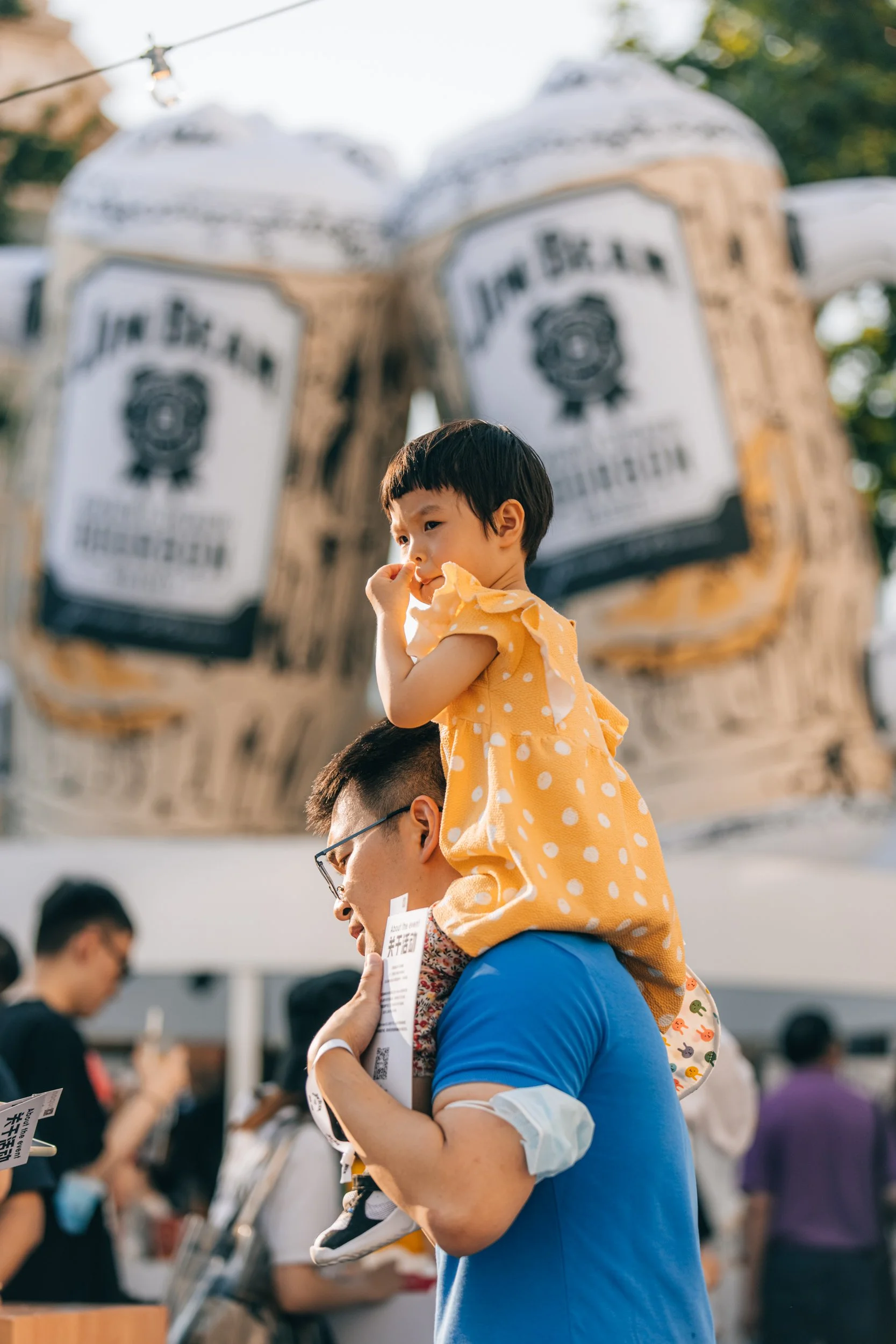 A young girl in a yellow dress with white polka dots is sitting on a man's shoulders at an outdoor event, with three large balloons behind them. The man is holding a white paper, and the scene is filled with other people and stalls in the background.