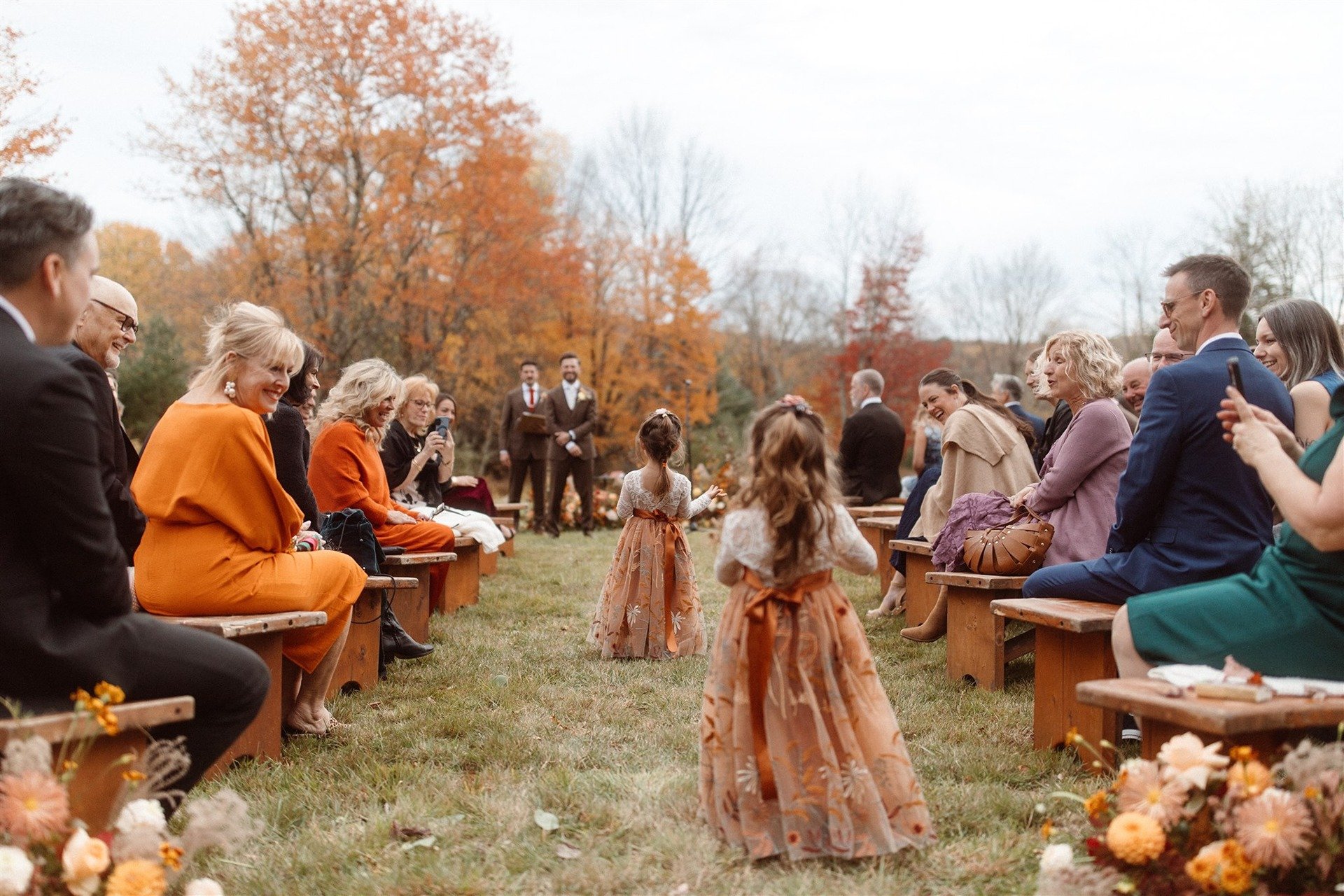 Two-Flower-Girls-In-Burnt-Orange-Fall-Dresses-Walking-Down-The-Aisle-In-An-Outdoor-Ceremony-At-Hemlock-Neversink-In-The-Hudson-Valley-New-York