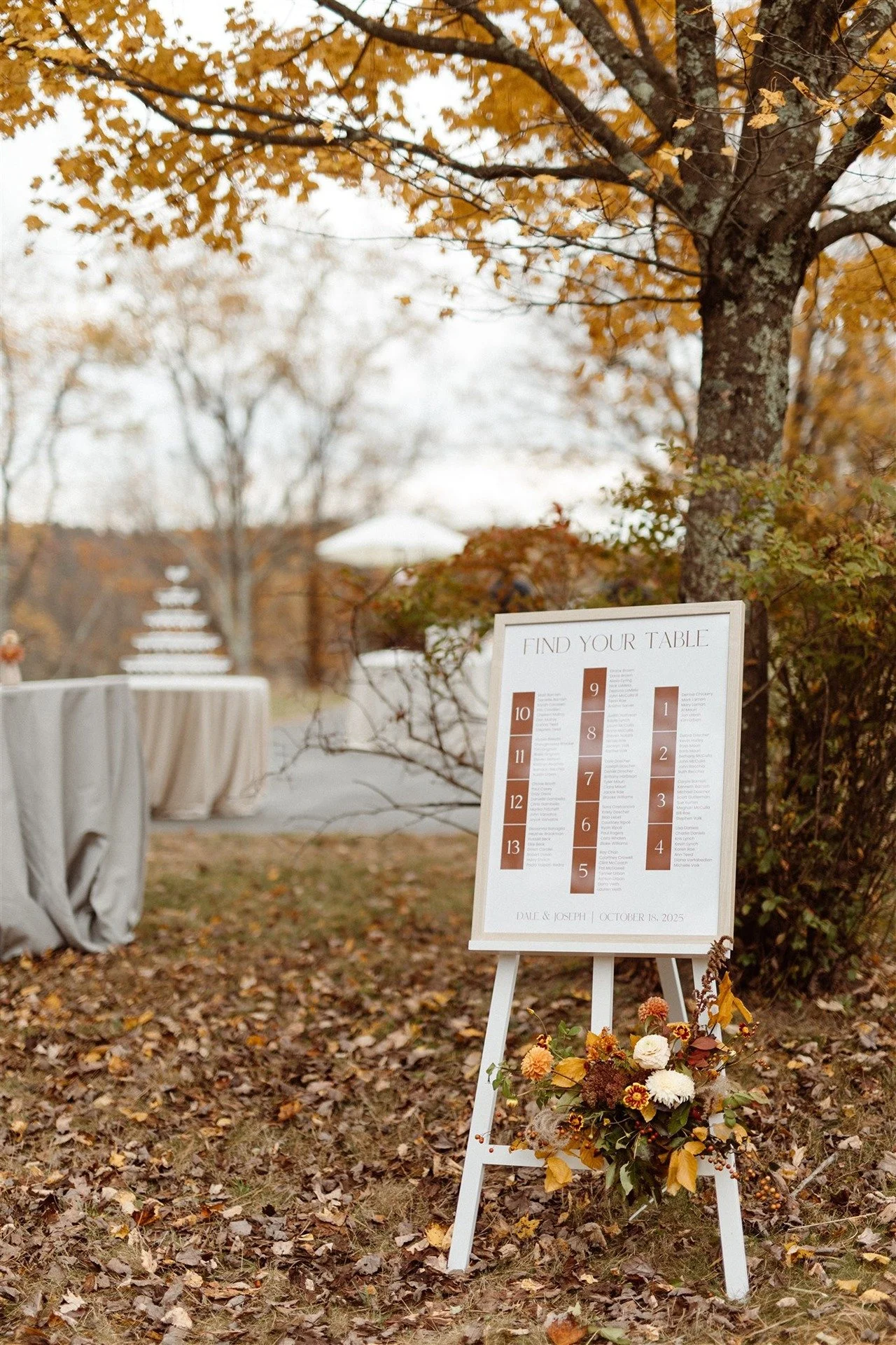 White-Wooden-Easel-With-Wedding-Seating-Chart-Set-In-Front-Of-A-Big-Maple-Tree-With-Champagne-Tower-In-The-Background
