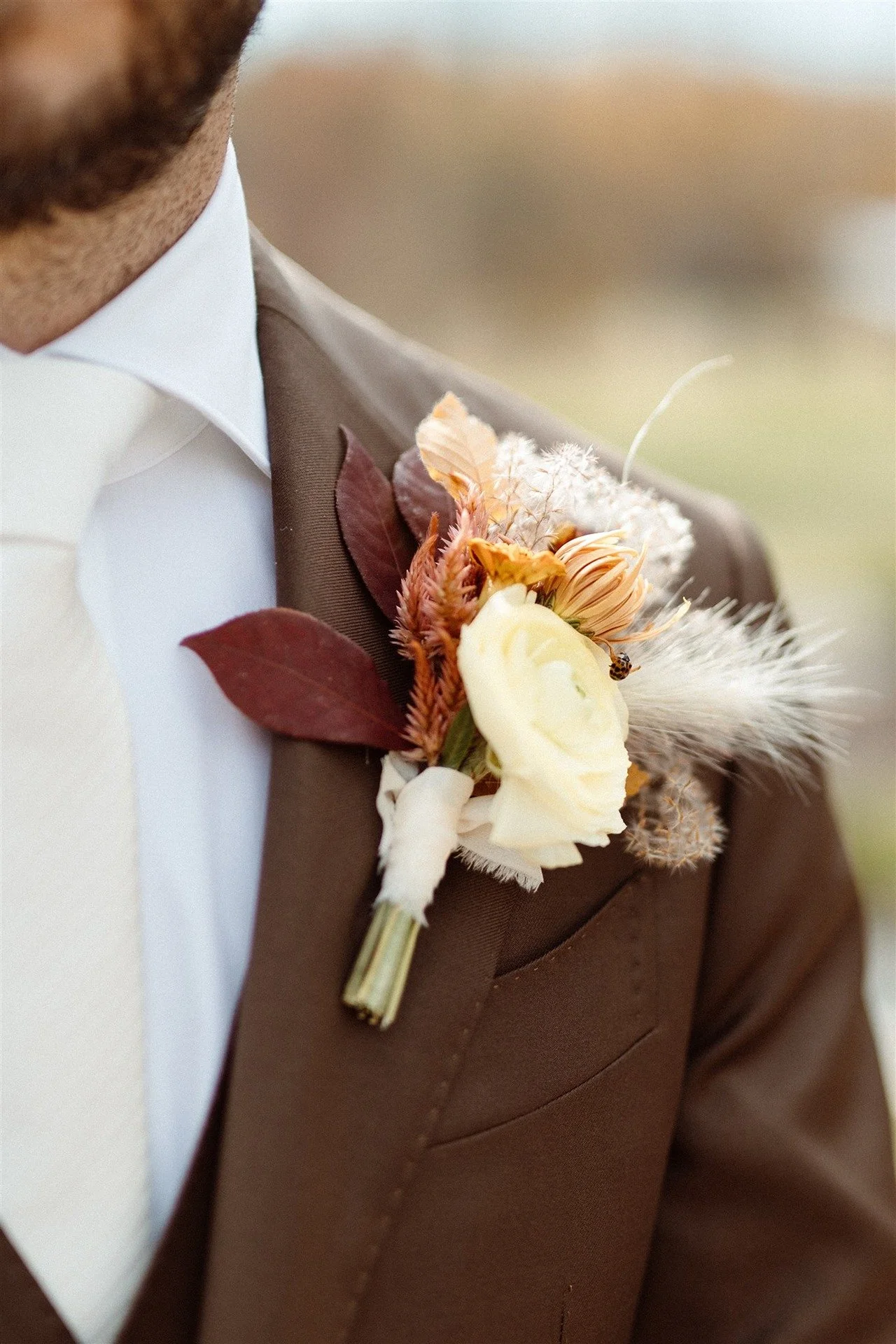 Elaborate-Fall-Boutonniere-With-Garden-Roses-Feathers-Amaranth-On-A-Brown-Suit-With-White-Tie