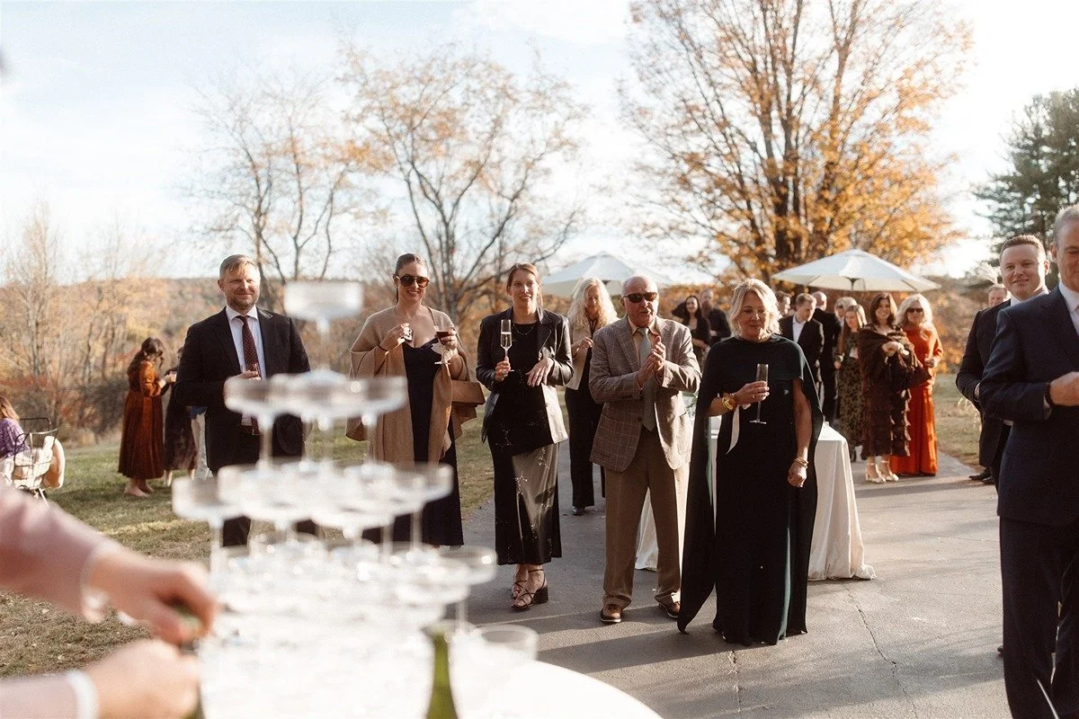 Cocktail-Hour-At-An-Outdoor-Wedding-In-The-Hudson-Valley-With-Champagne-Tower-Being-Set-Up-And-Guests-Admiring-In-The-Background
