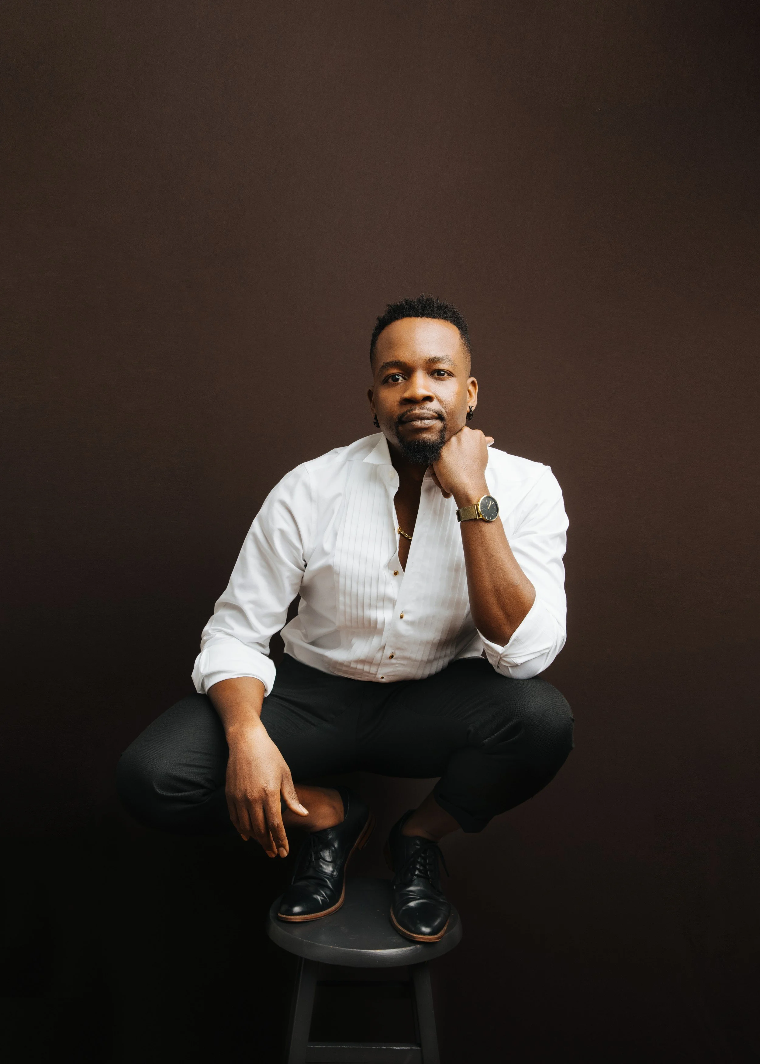 Portrait of Nigerian-American baritone Babatunde Akinboboye in a white tuxedo shirt, crouching on a stool.