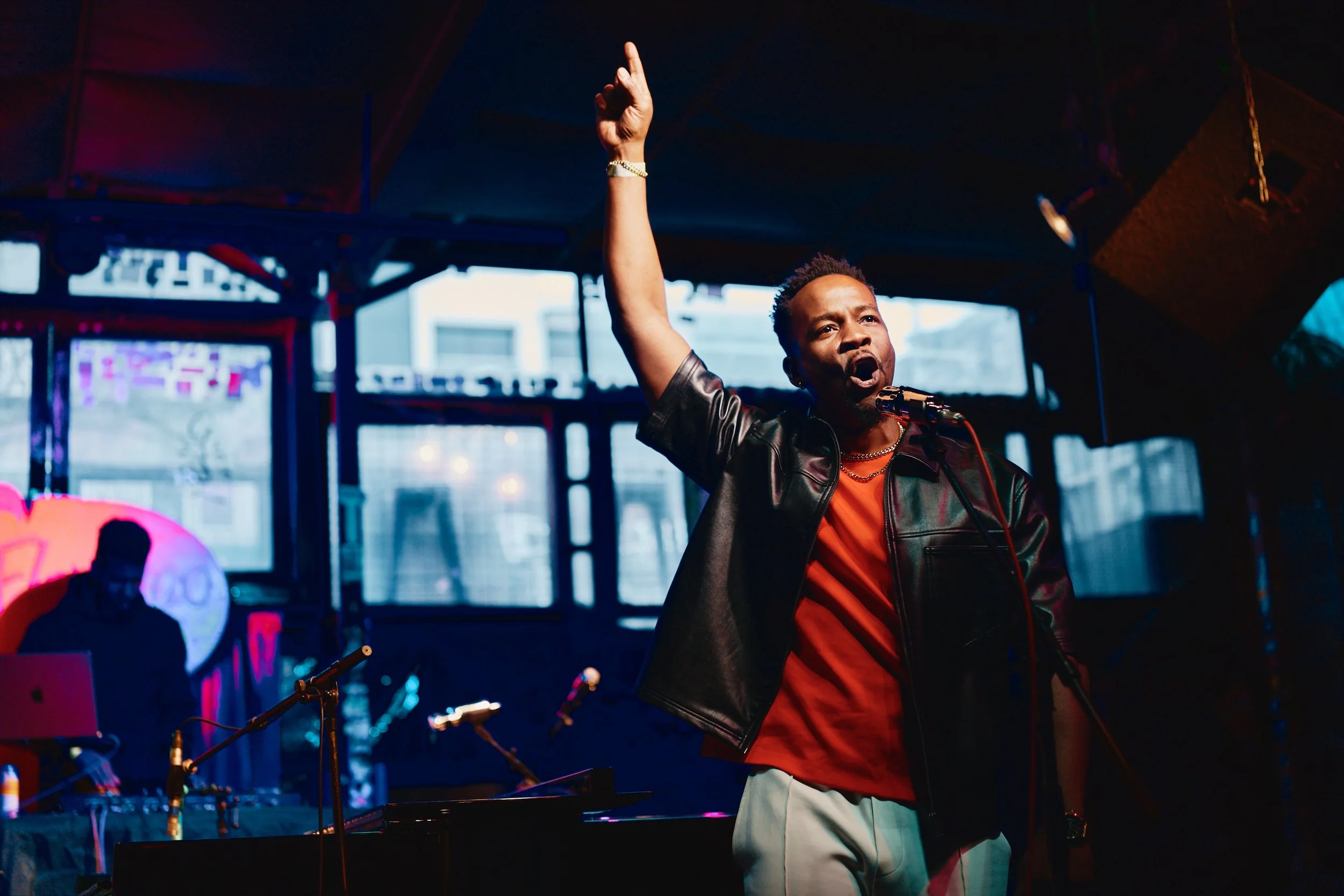 Babatunde Akinboboye performing HipHopera onstage in a black leather jacket and red shirt.
