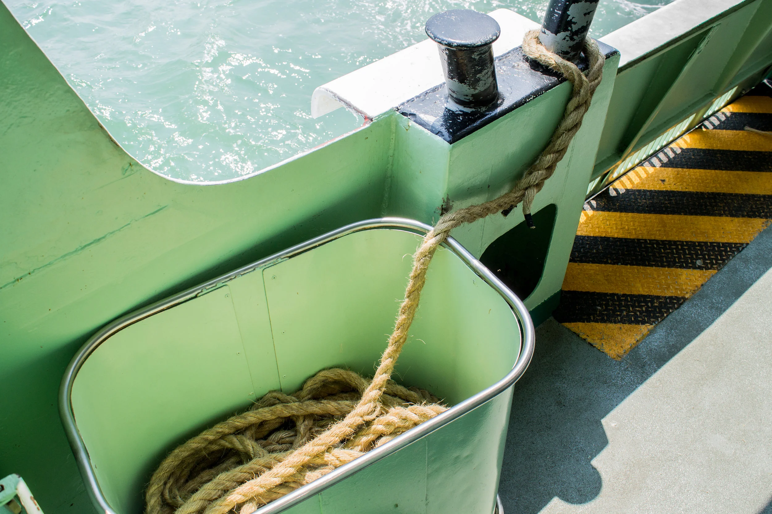 Close-up of a green boat dock with black and yellow safety markings, a metal basket with coiled rope, black bollard, and cleat with tied rope.